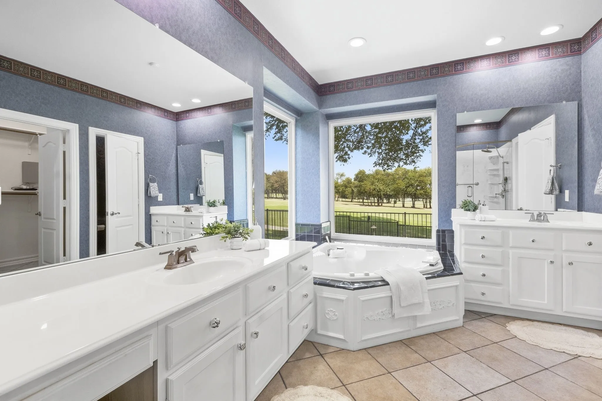 Primary Bathroom featuring a picturesque view of nature,  jetted soaking tub, walk-in shower with glass enclosure, dual vanities with white cabinetry, and tile flooring