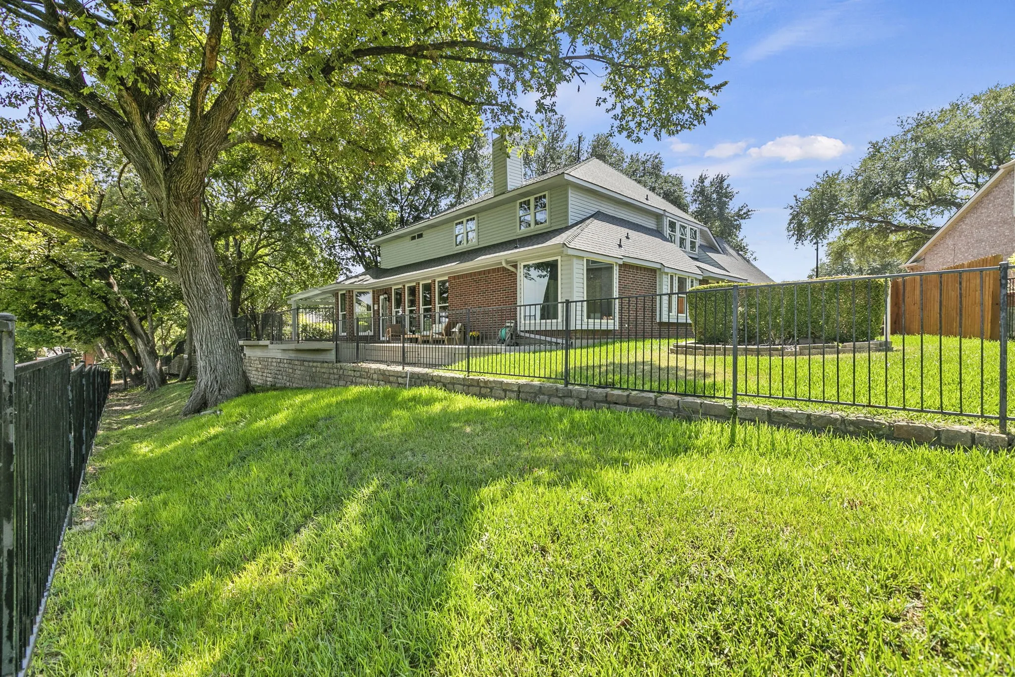 Exterior with an expansive lawn, mature trees, a wrought iron fence, a brick retaining wall, gutters with leaf guards, sprinkler system, Pergolas, Anderson windows