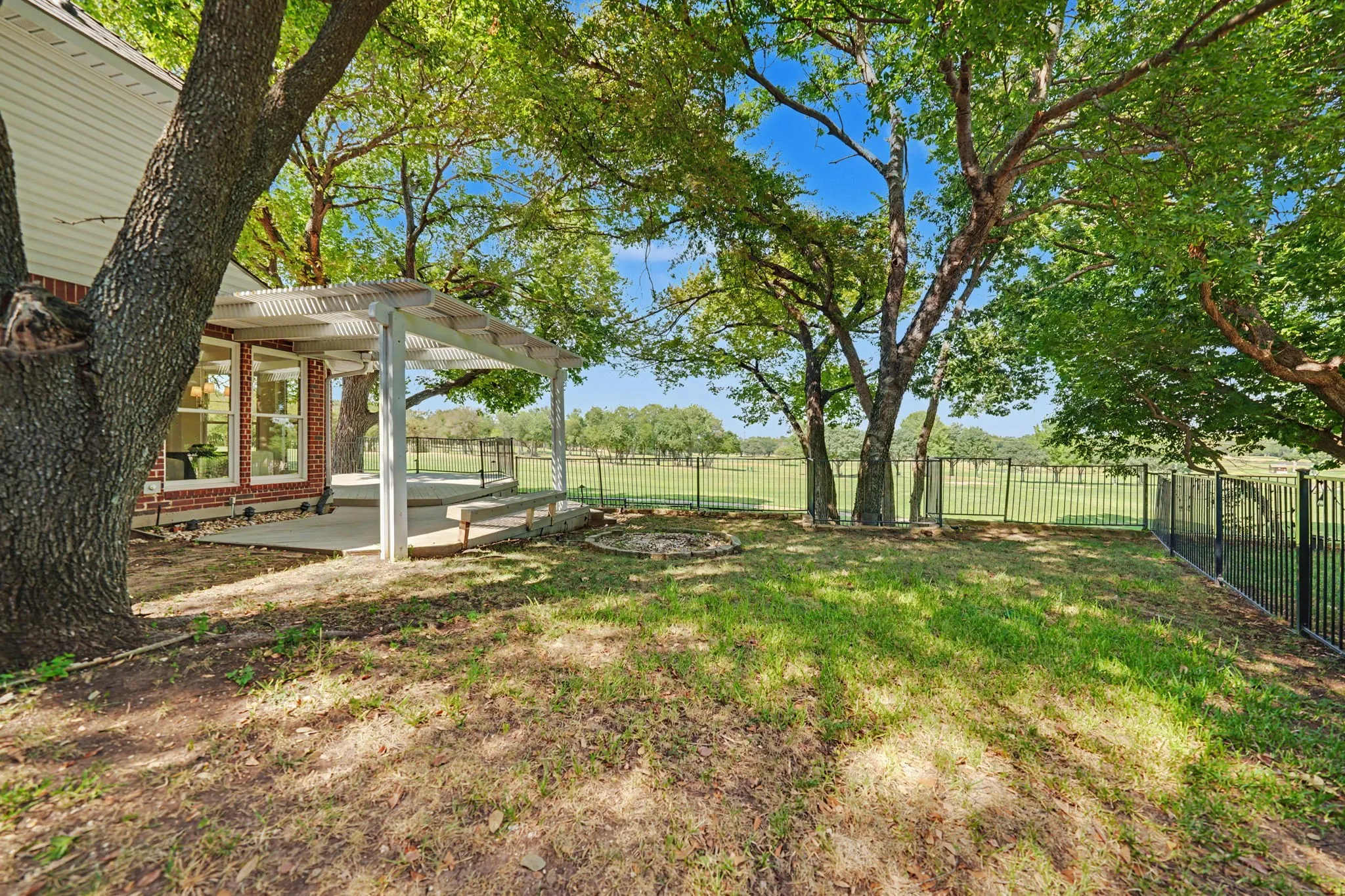 Expansive backyard with a pergola, mature trees,  a nicely stained wood gate up front and a metal fence lining the sides and back of the property.