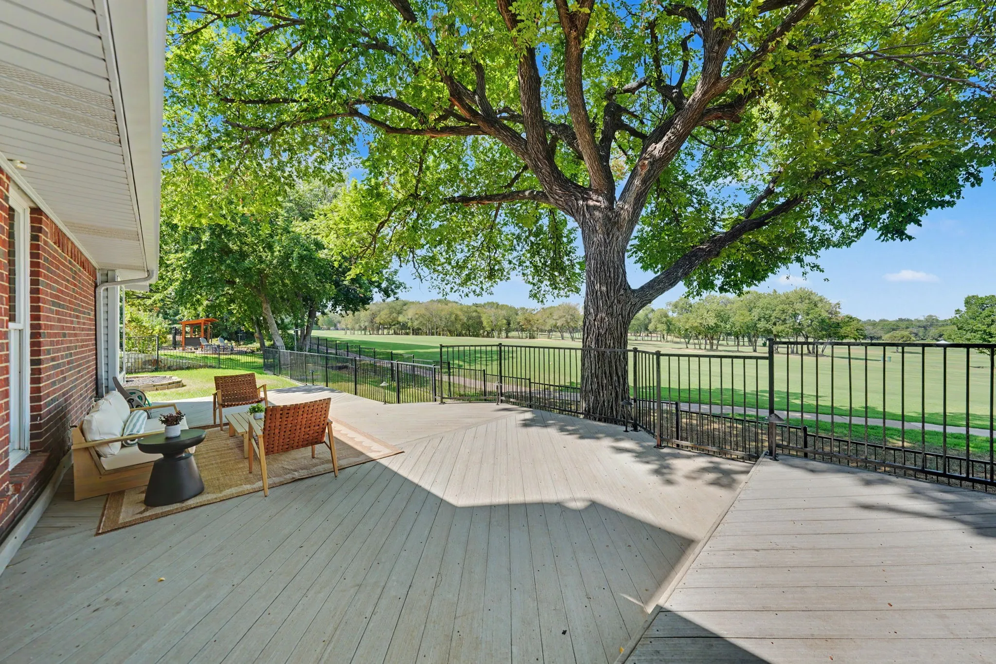 Expansive outdoor deck with a mature tree, bordered by a black metal fence, overlooking a manicured greens.