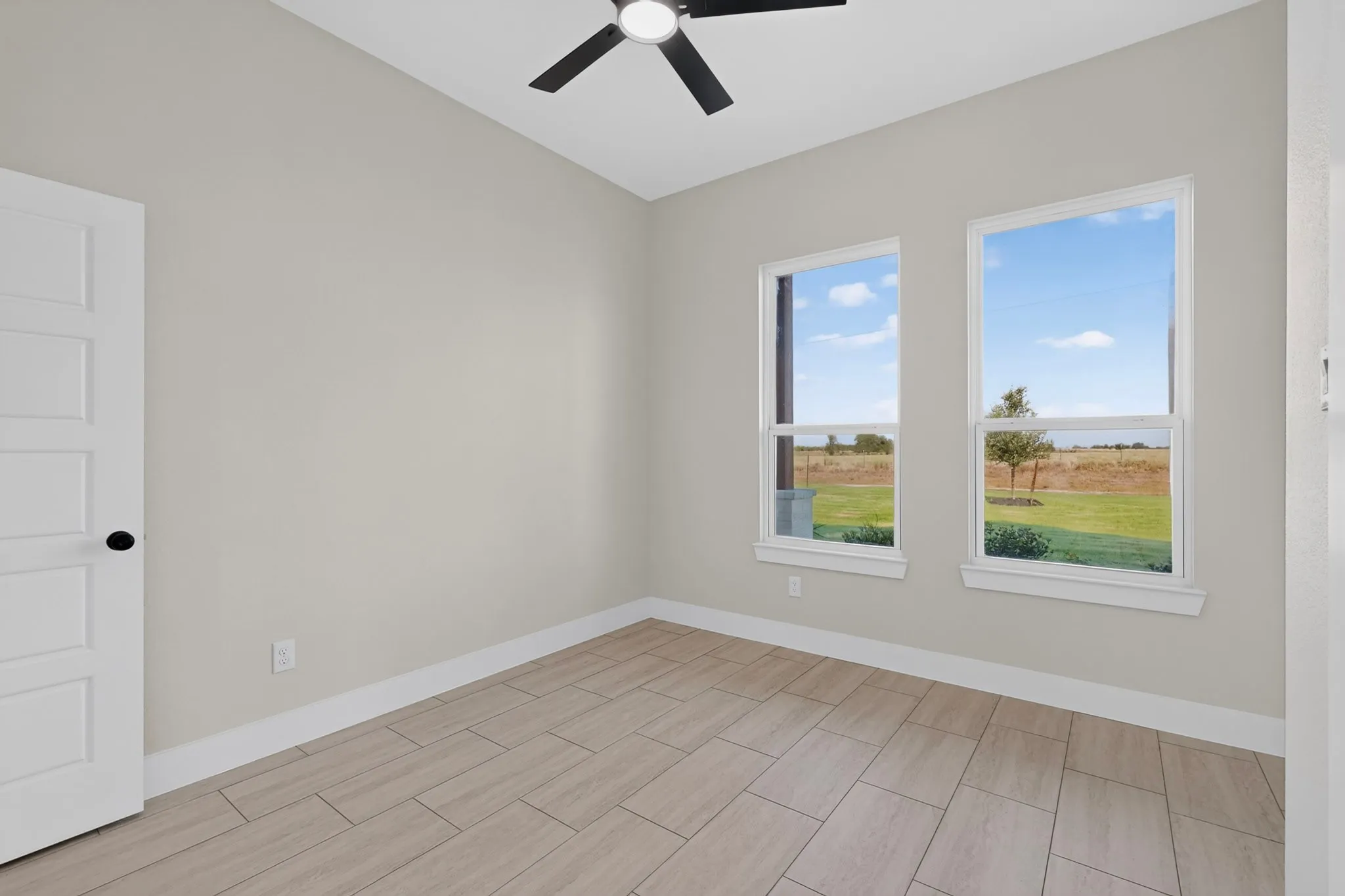 Room with wood-finish tile flooring, two large windows, a black ceiling fan, and a white paneled door with a black doorknob