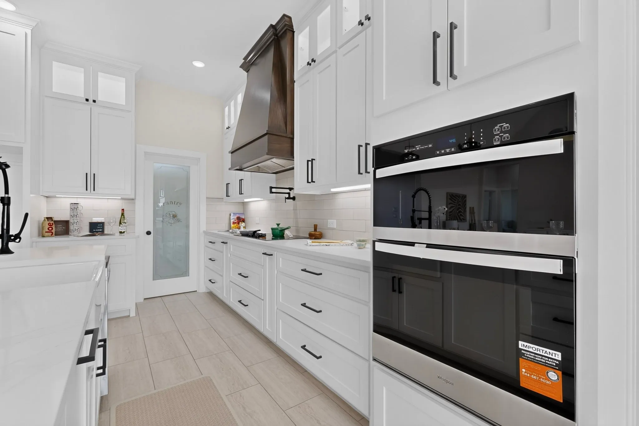 White shaker cabinetry with dark hardware, integrated double ovens, wood-finish hood vent, tile flooring, and a frosted glass pantry door