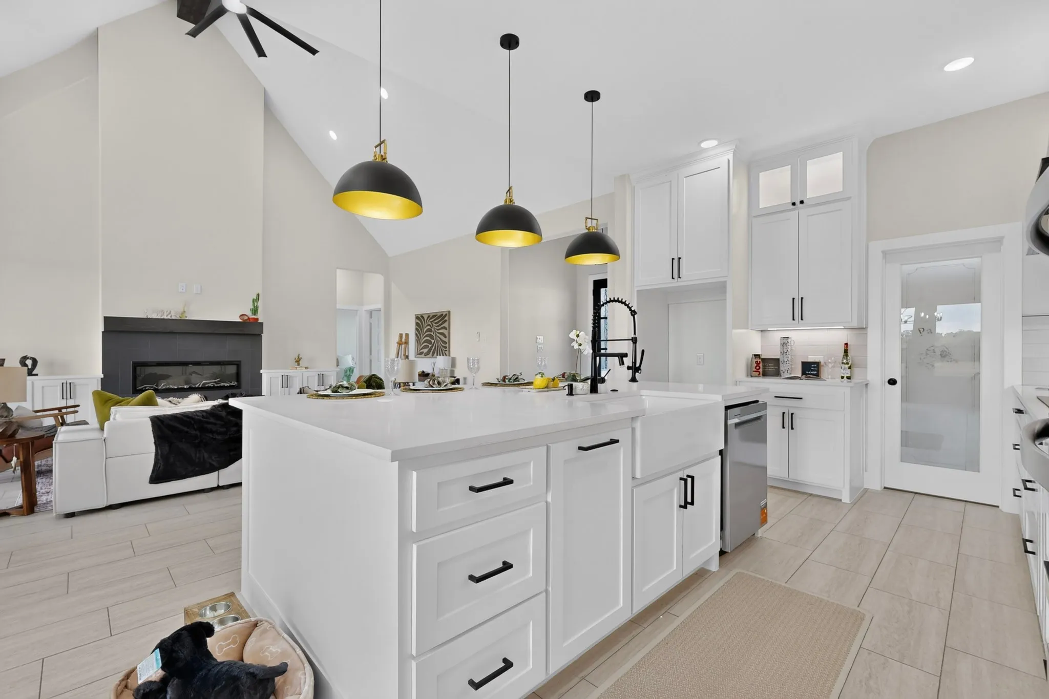 Modern kitchen featuring white cabinetry with black hardware, a large waterfall-edge island with storage drawers, stainless steel appliances, pendant lighting with brass-finished domes, quartz countertops, and light wood-finish flooring throughout the open-concept space