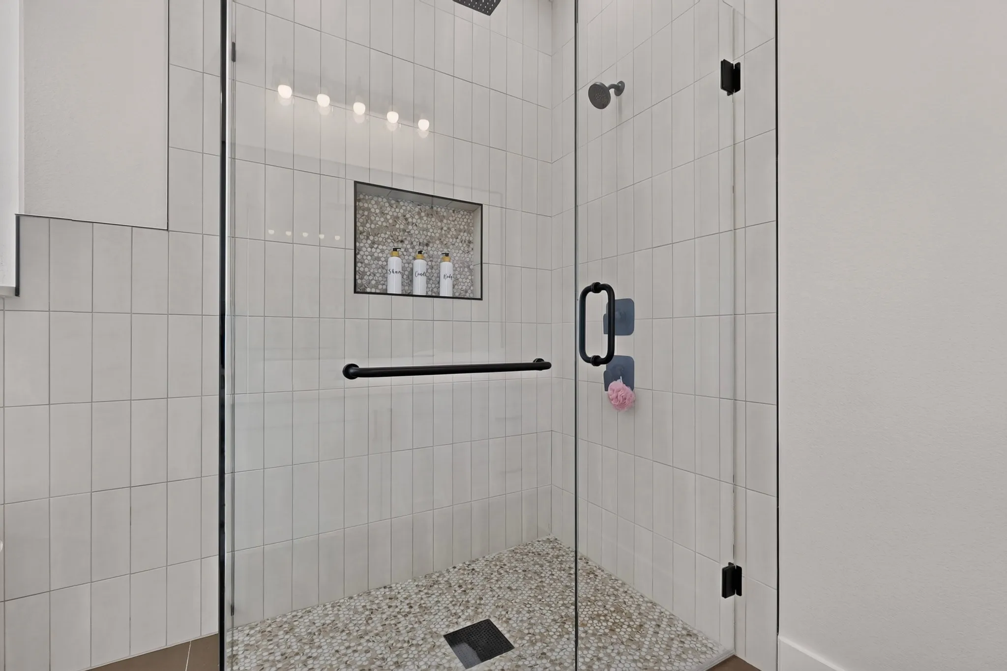 Frameless glass shower enclosure with matte black hardware, vertical-set white tile surround, integrated shower niche with mosaic tile, and a pebble-style shower floor