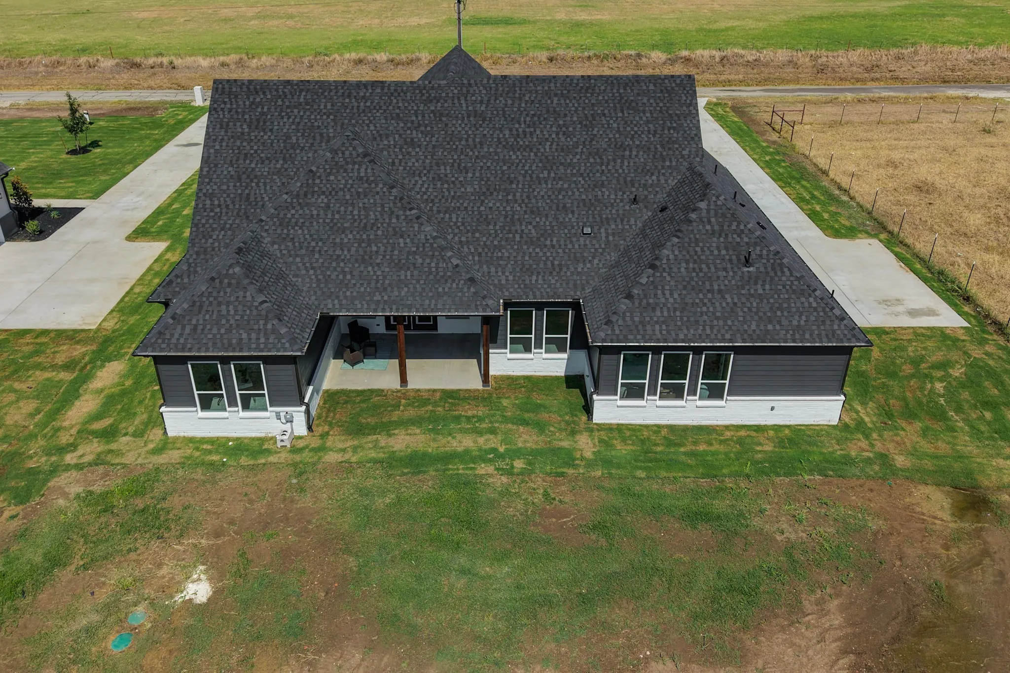 Expansive dark shingle roof, covered patio with wood columns, concrete patios, and mature green landscaping