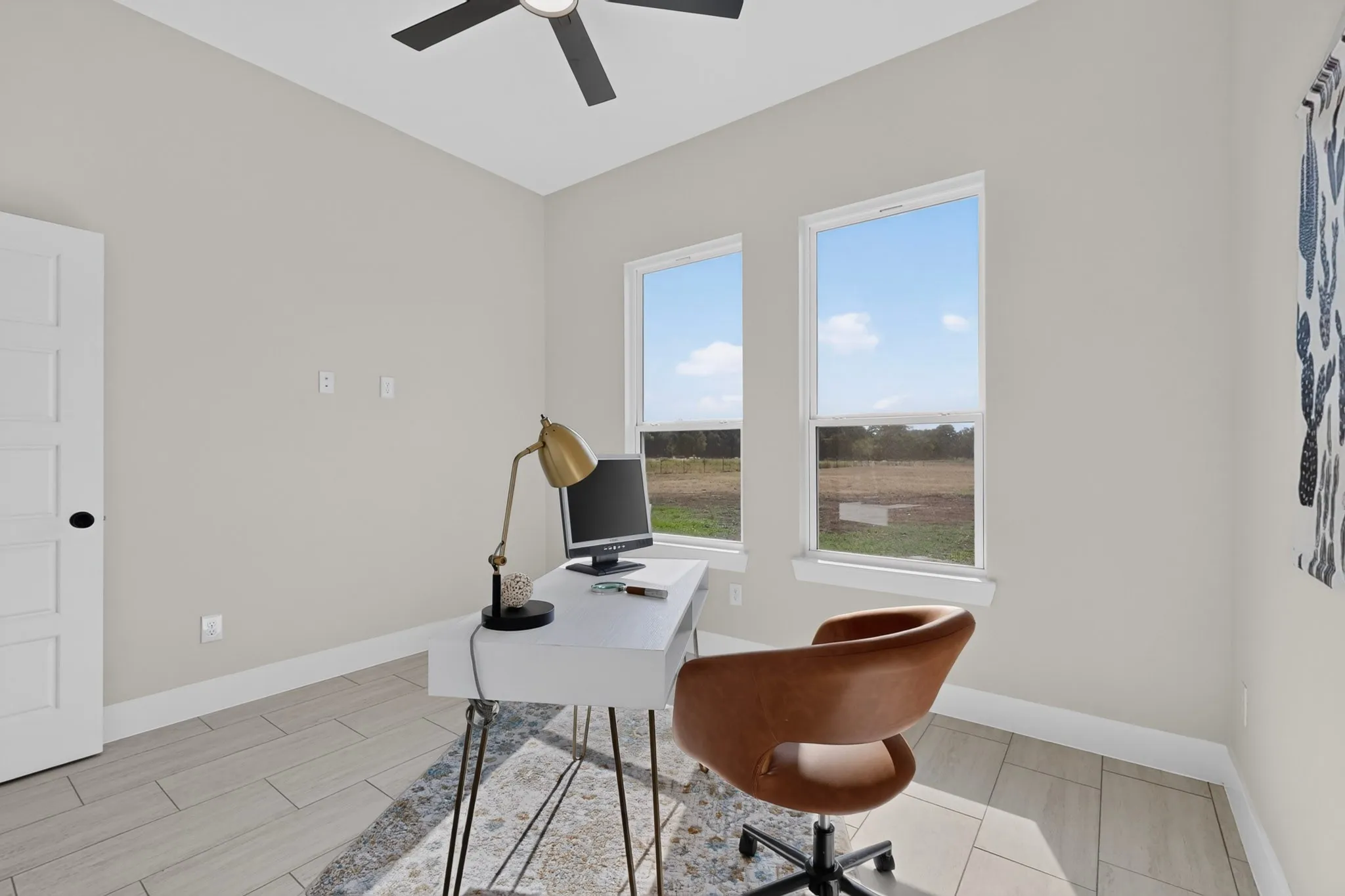 Bright home office featuring expansive windows overlooking pastoral landscape, wood-finish flooring, ceiling fan with remote control, and a curved brown leather desk chair