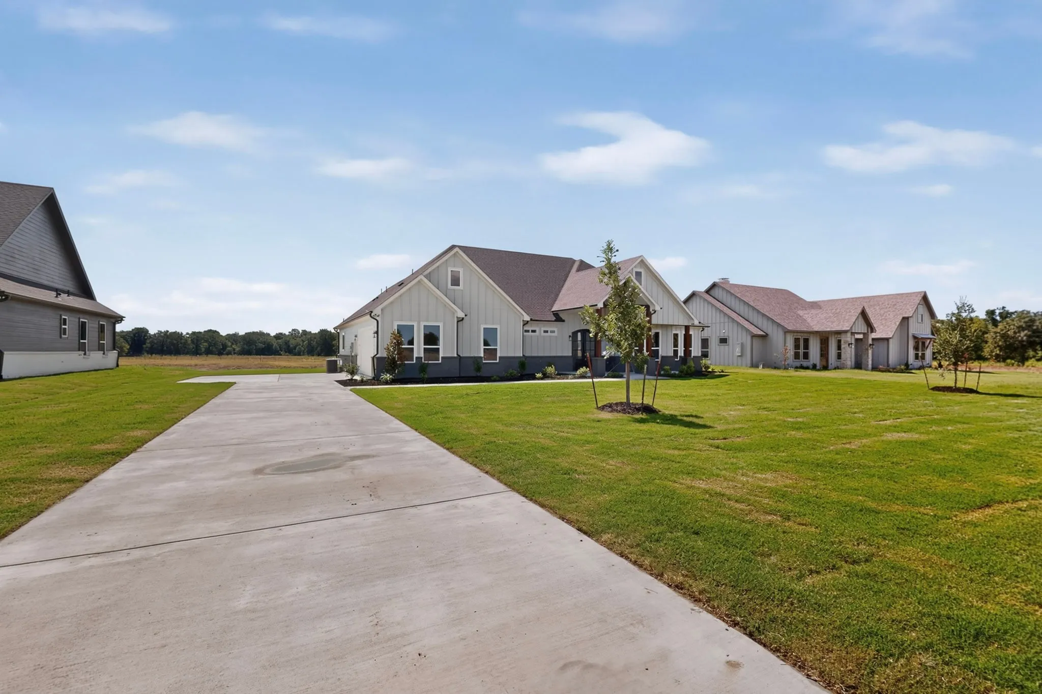 Concrete drive leading to a light-colored siding residence with a dark roof, surrounded by expansive green lawn and distant trees