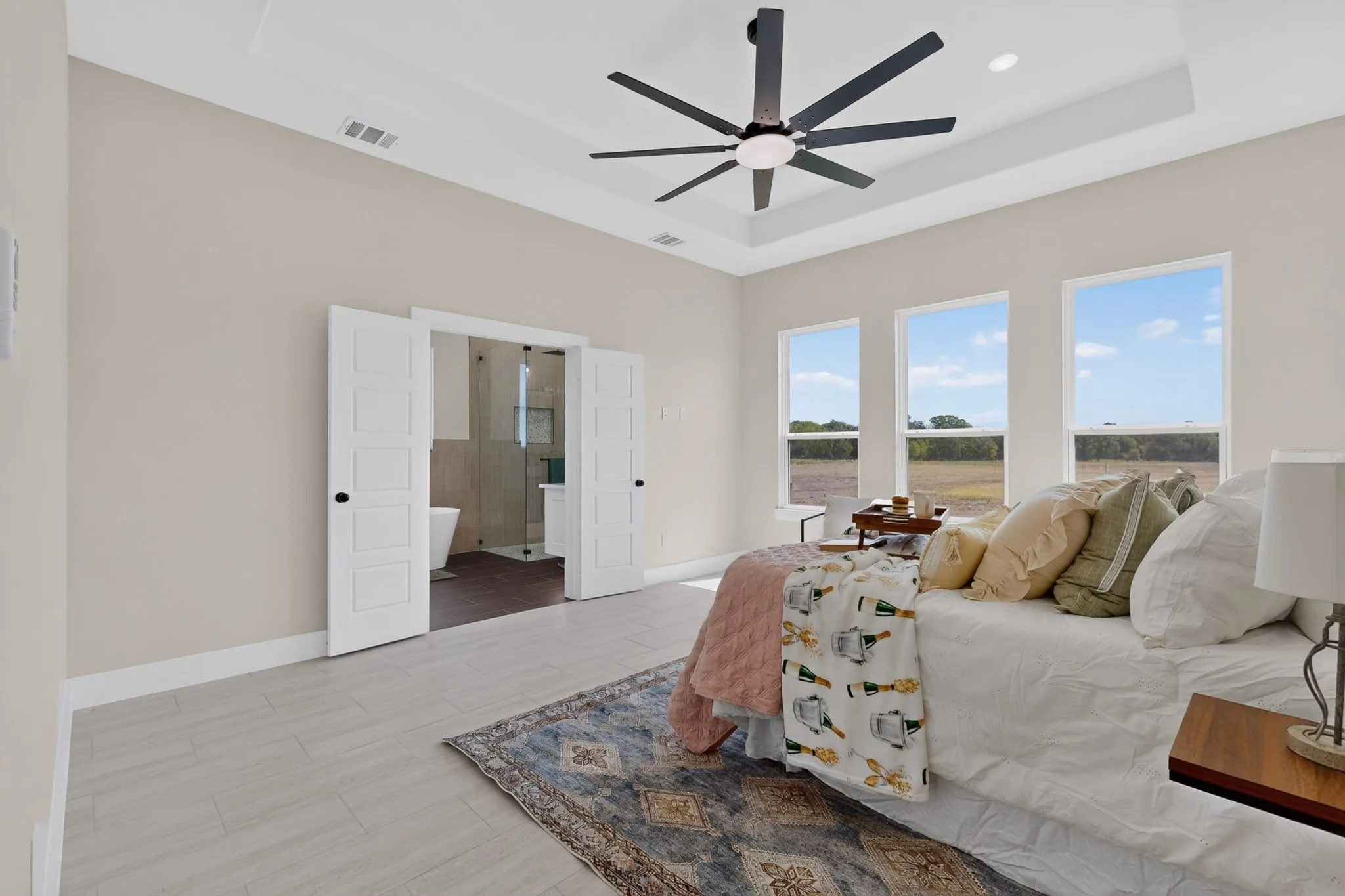 Tray ceiling with a modern ceiling fan, three large windows, light-colored wood-finish flooring, and double paneled doors opening to a bathroom