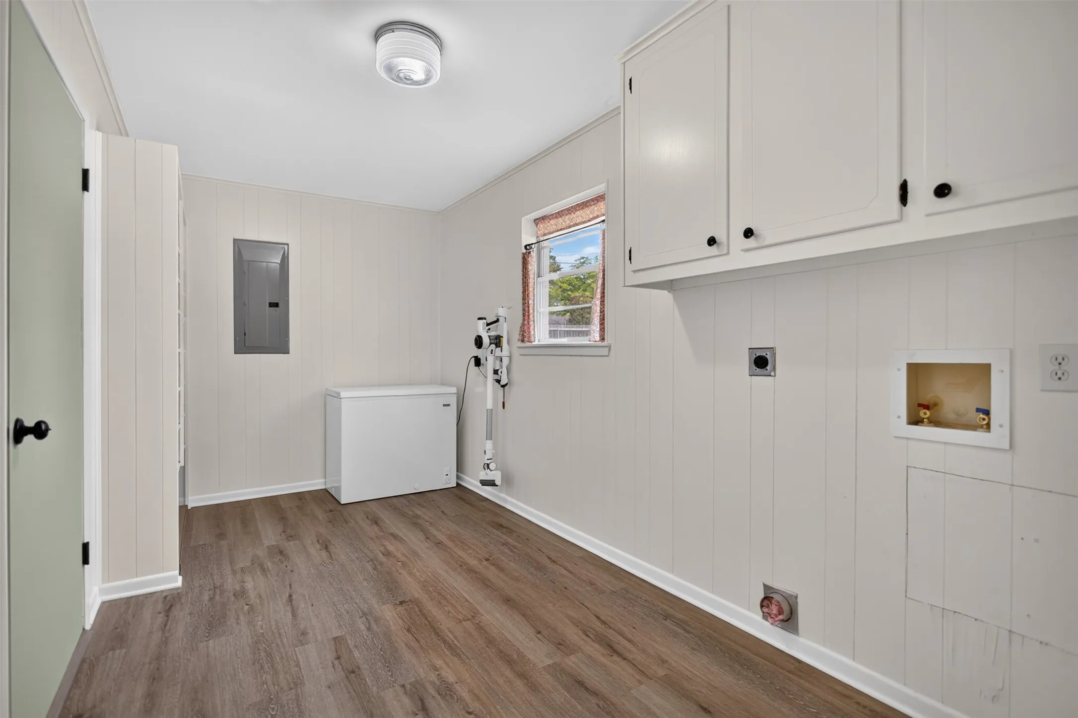 Wood-finish flooring, white paneled walls, recessed ceiling light fixture, white upper cabinetry, and laundry hookups