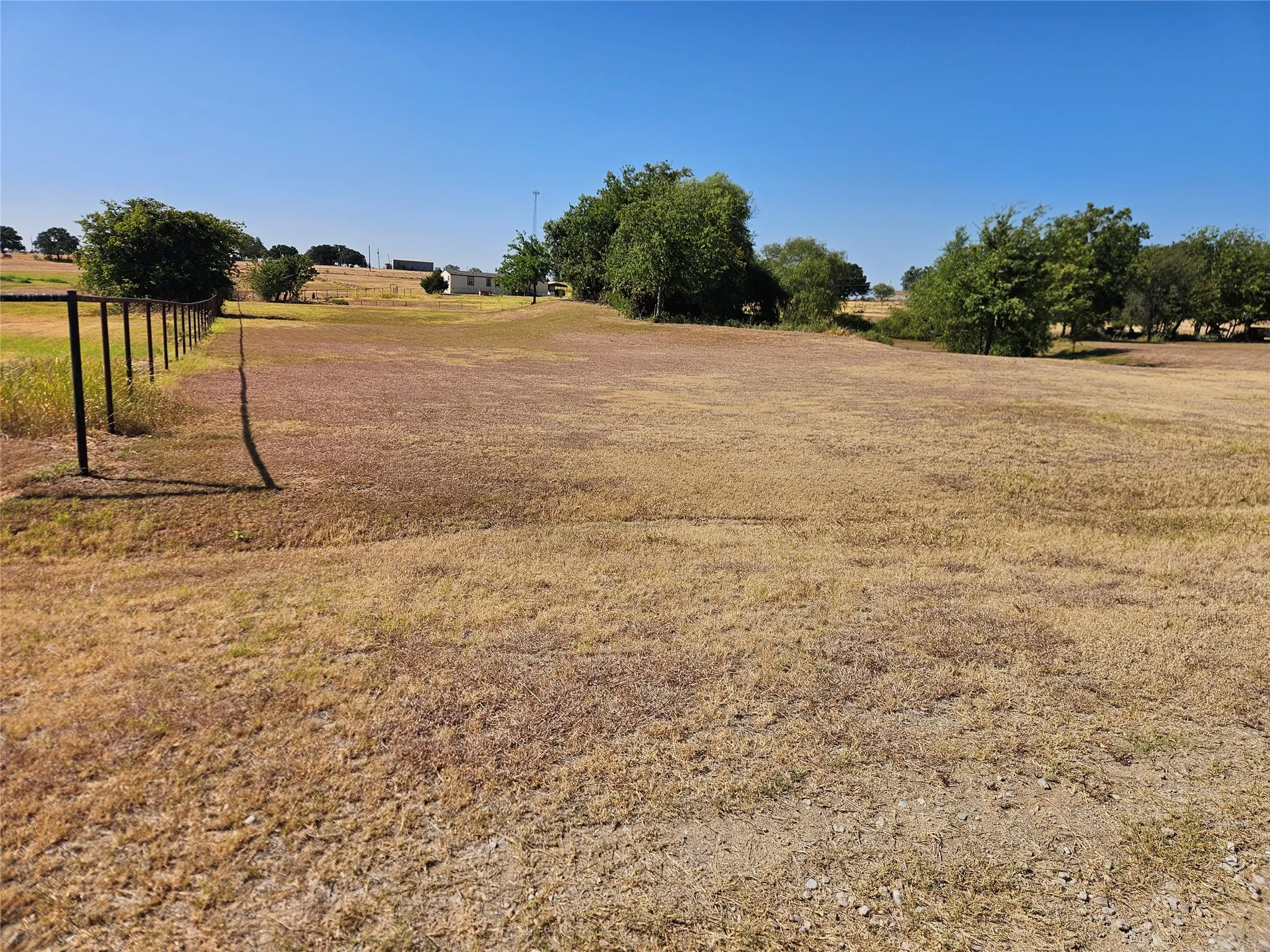 Expansive open land area with a clear sky backdrop, featuring a partial metal fence line and distant green foliage.