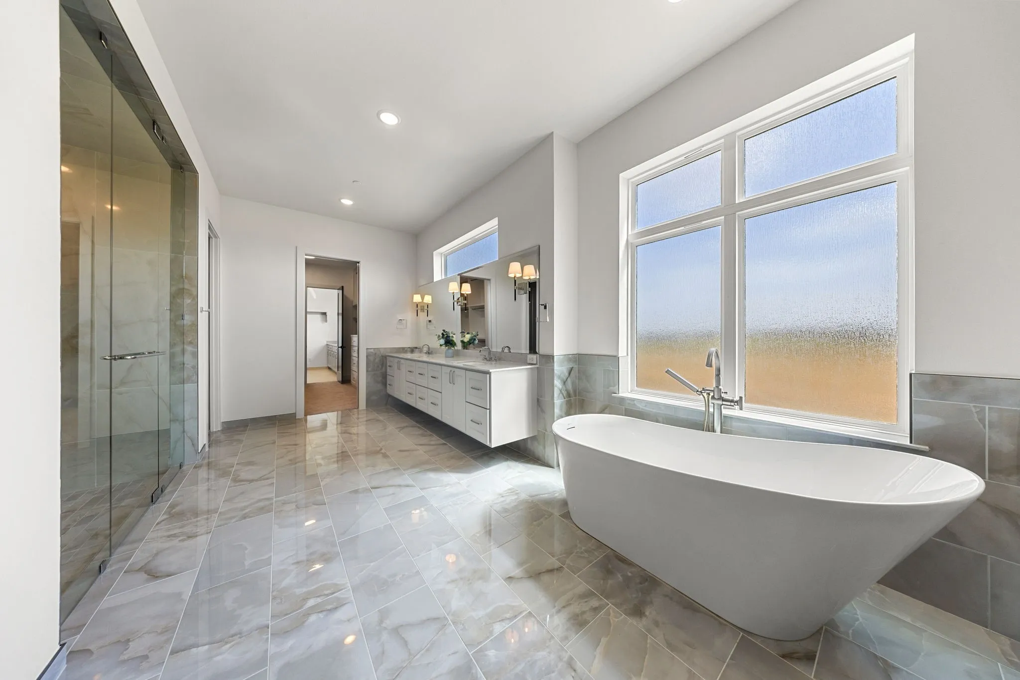 Freestanding soaking tub, large window, dual vanity with undermount sinks, glass-enclosed shower, and tile flooring