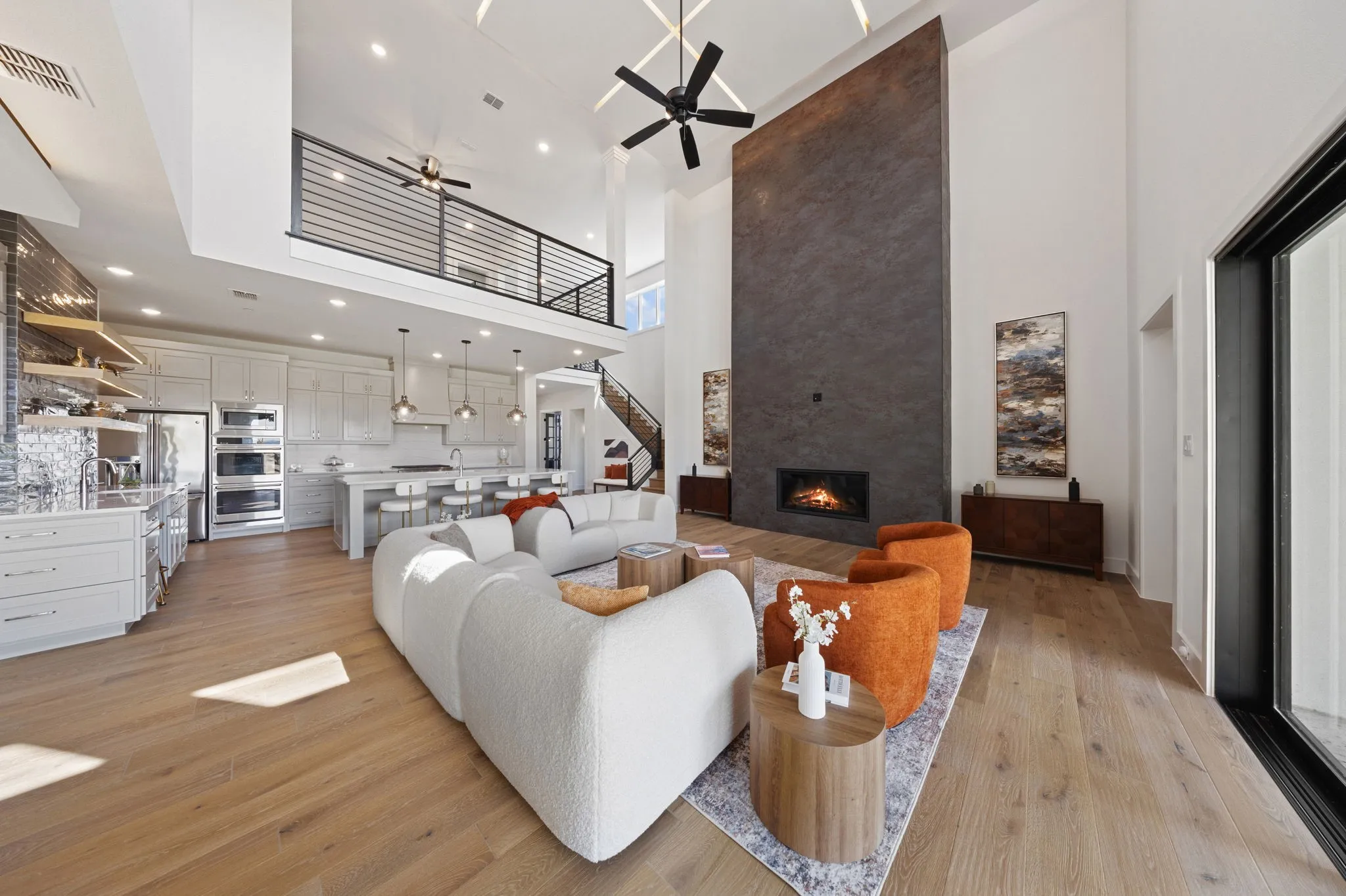 Open-concept living area with wood-finish flooring, a two-story fireplace with a built-in firebox, and a gourmet kitchen featuring white cabinetry, an island with seating, and stainless steel appliances