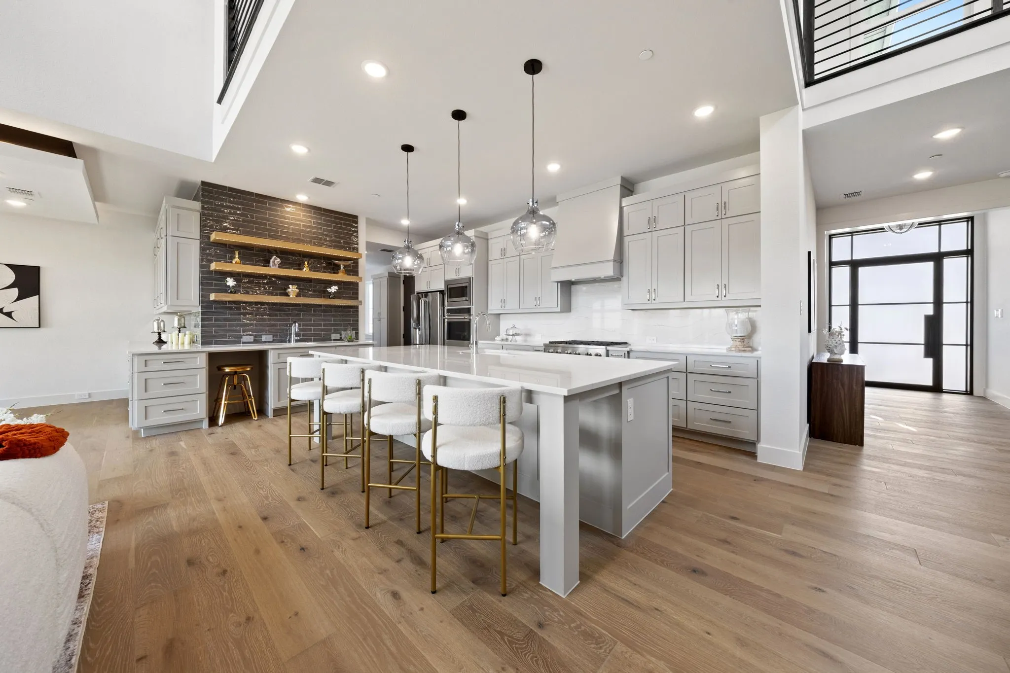 Open-concept kitchen featuring light wood-finish flooring, a central island with countertop seating, light gray cabinetry, built-in stainless steel appliances, and a brick-accented wet bar with open shelving