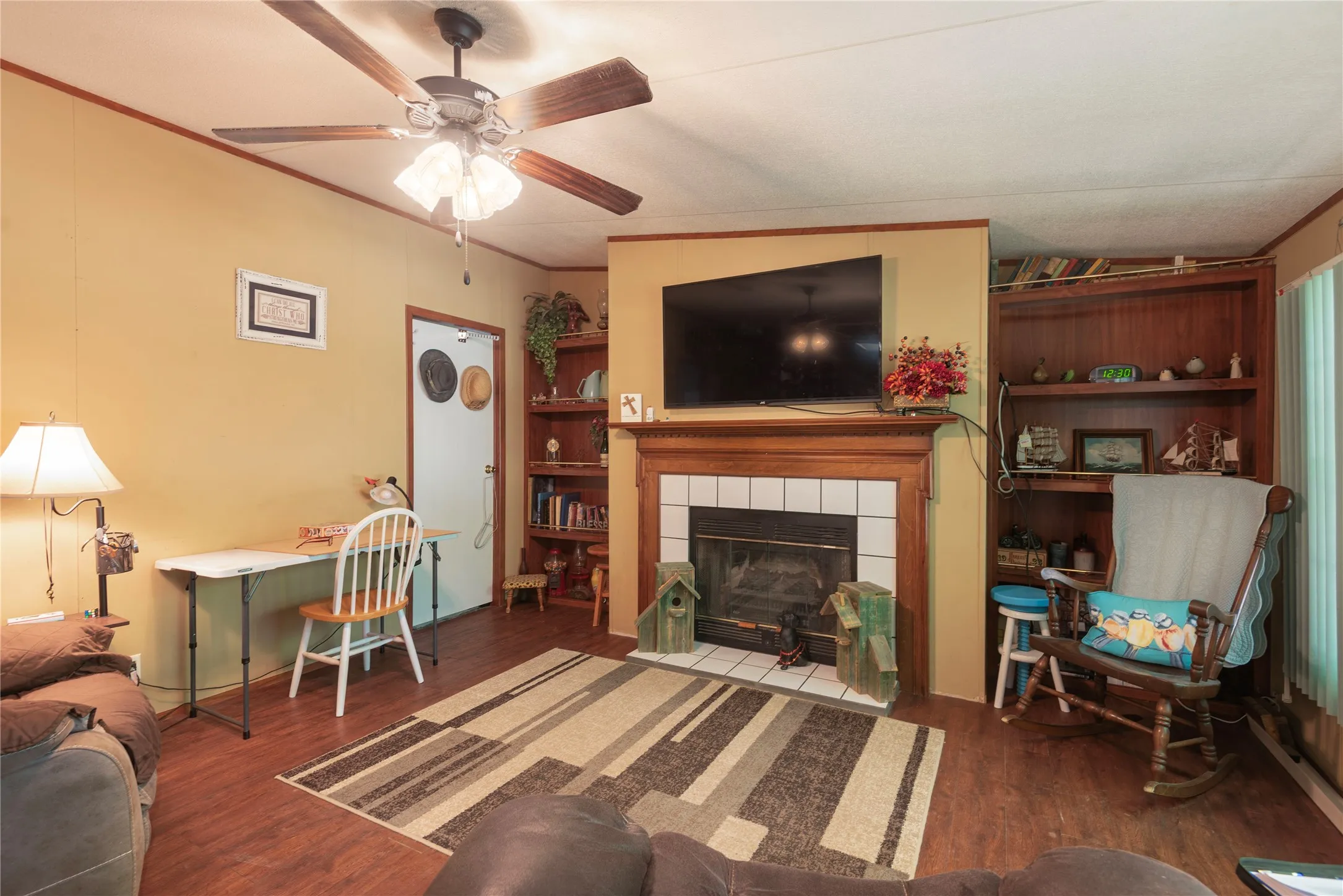 Fireplace with tile surround and wood mantel, wood-finish flooring, ceiling fan with integrated lighting, built-in shelving, and a door with trim
