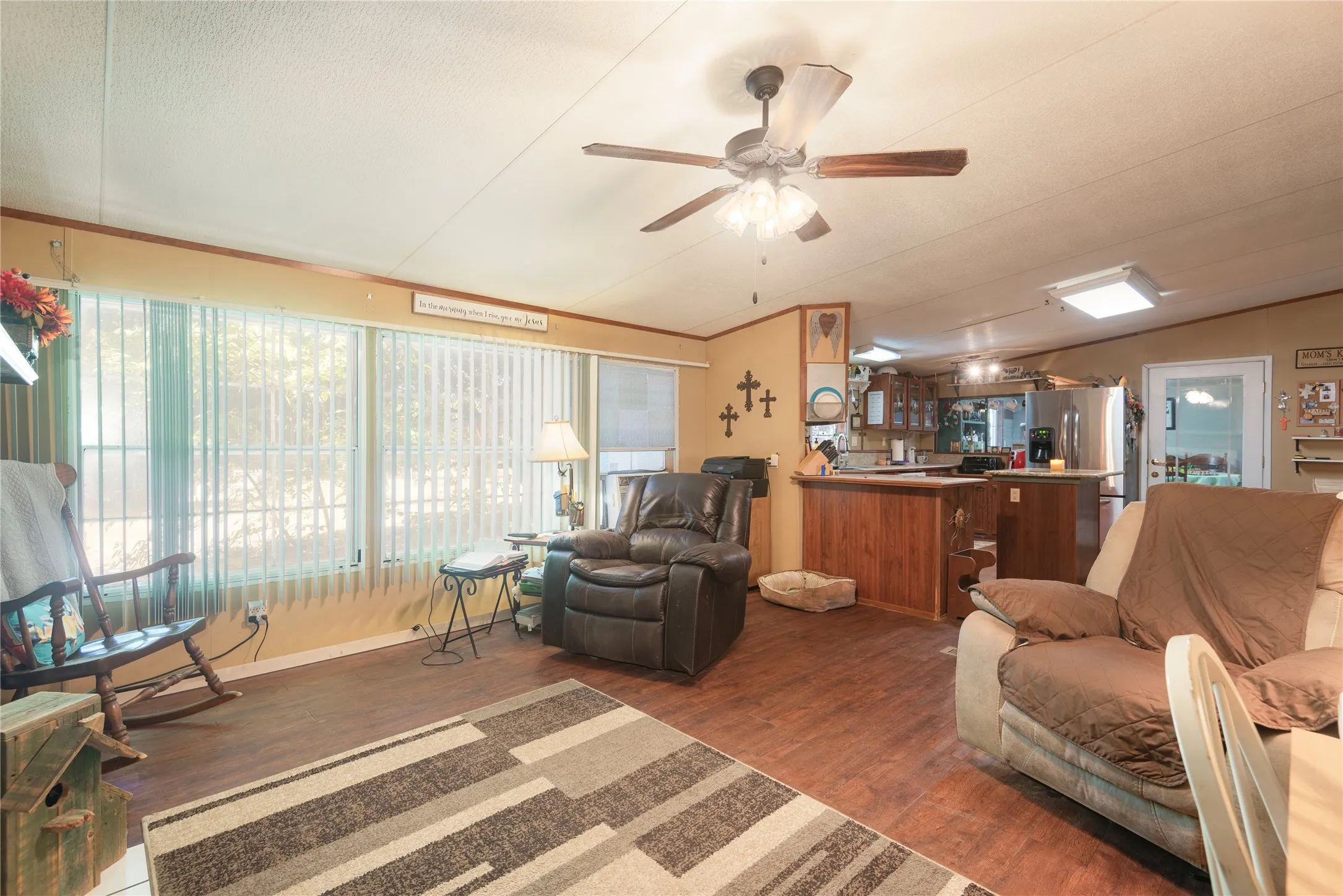 Wood-finish flooring throughout, multiple windows with vertical blinds, ceiling fan, and an open-concept layout leading to the kitchen area