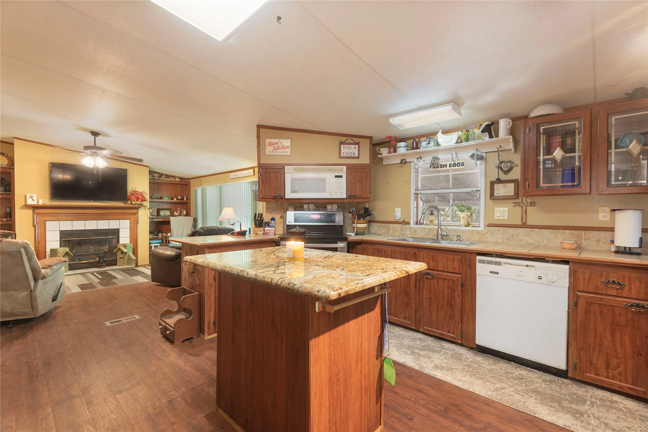 Open-concept kitchen and living area featuring a fireplace with a white mantel, wood-finish flooring, a kitchen island with a stone-finish countertop, and wood cabinetry