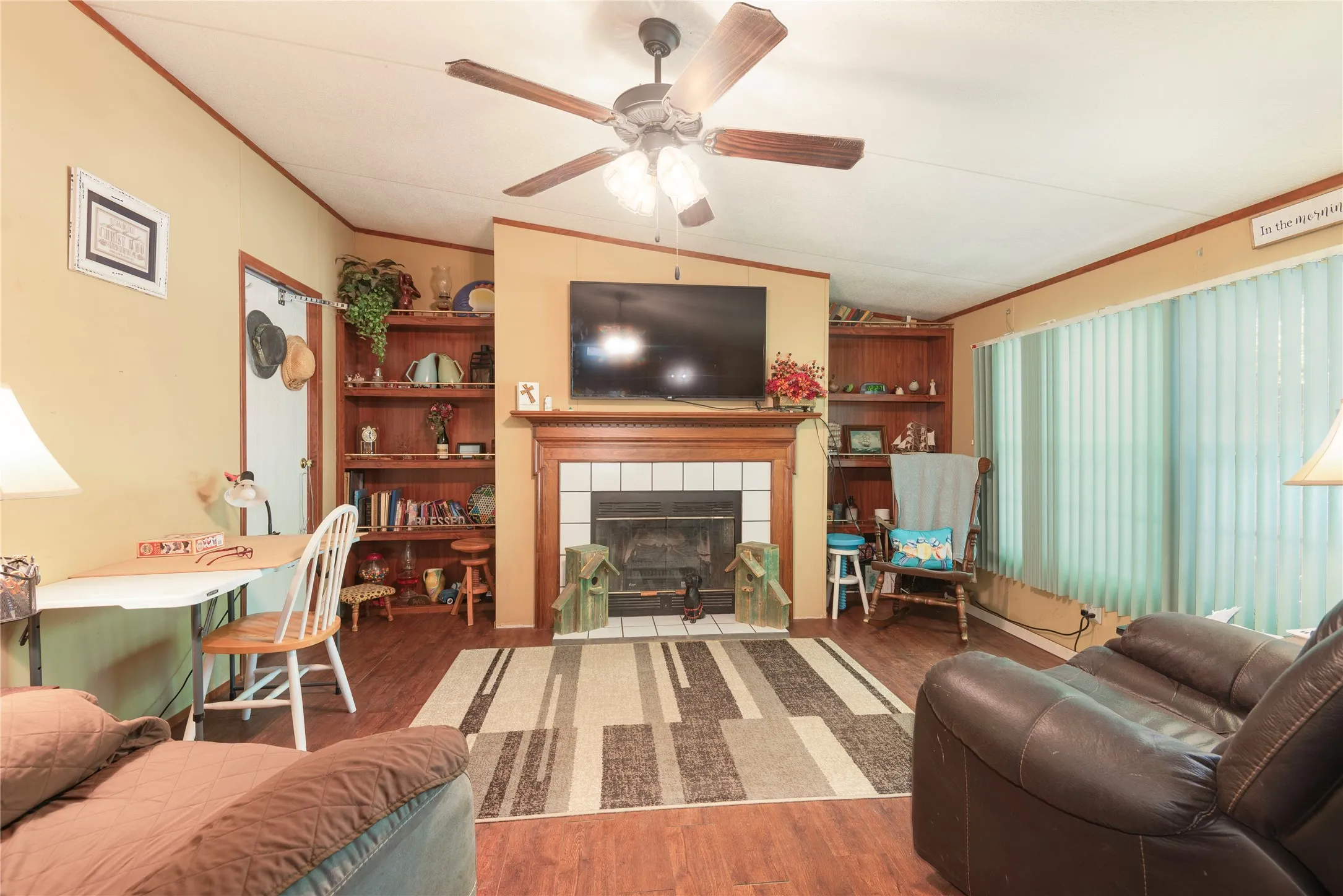 Wood-finish flooring throughout, a ceiling fan with integrated lighting, a fireplace with a tile surround and wood mantel, built-in shelving, and vertical window blinds