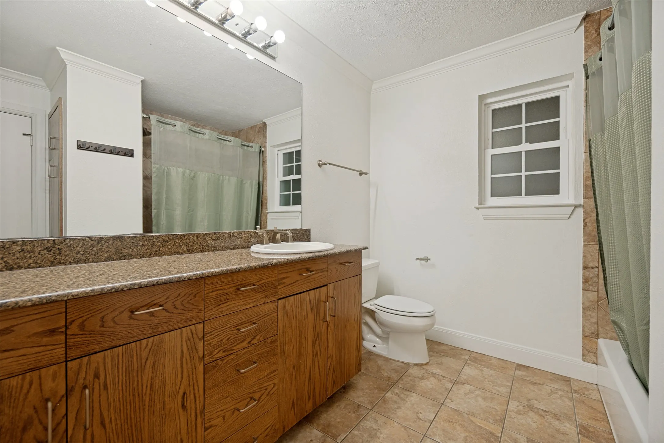 Wood-finish vanity with an integrated sink, granite-style countertop, large wall-mounted mirror, ceramic tile flooring, and a window providing natural illumination
