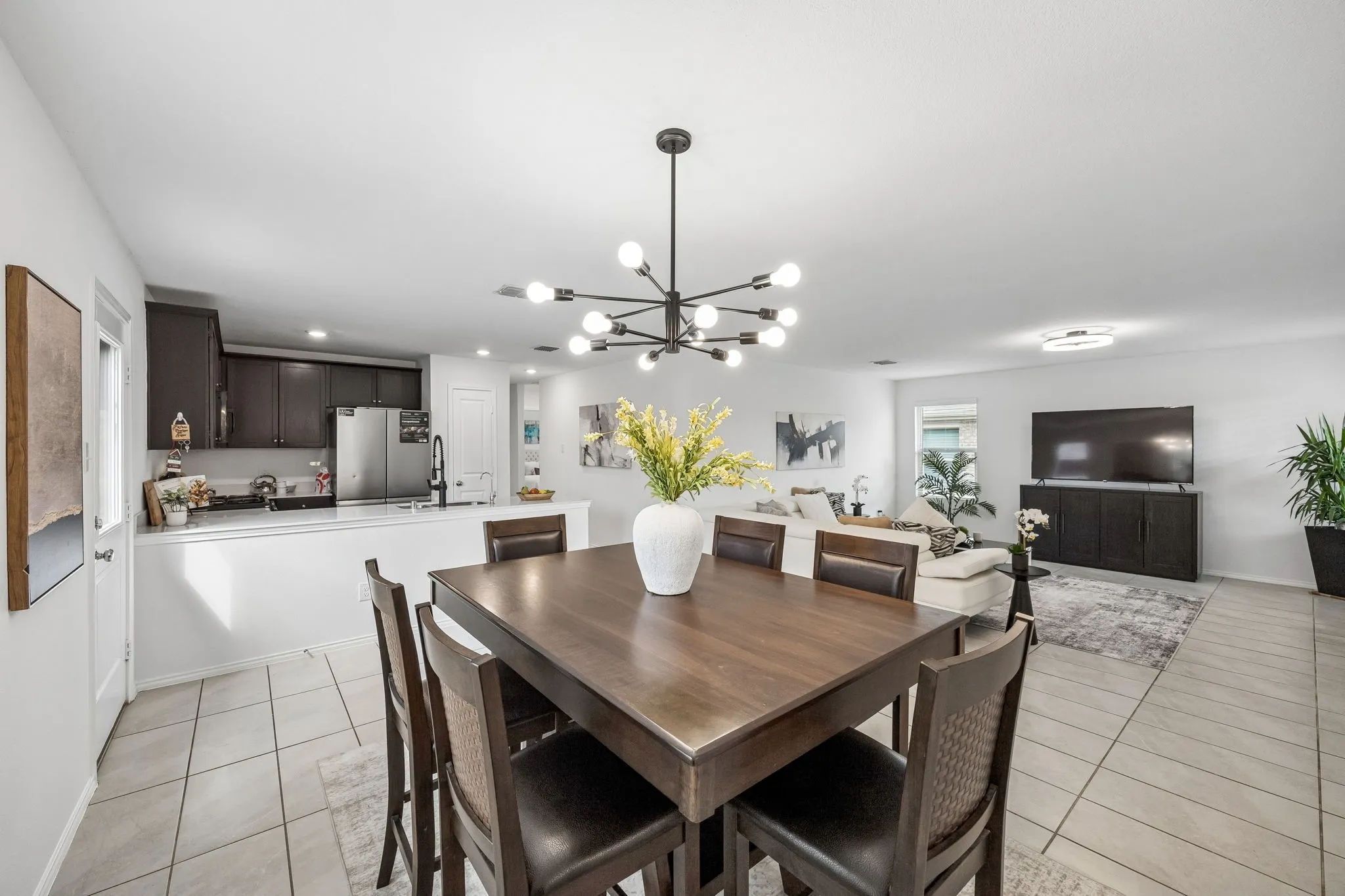 Open-concept living space featuring a dining area with a contemporary chandelier, a kitchen with dark cabinetry and stainless steel appliances, and a tiled floor throughout