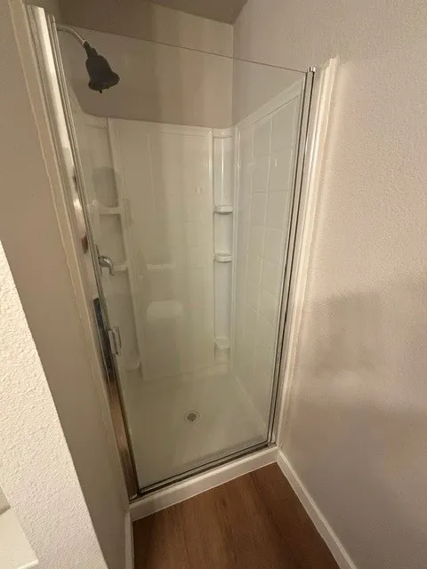 Walk-in shower with clear glass enclosure, white surround, and wood-finish flooring