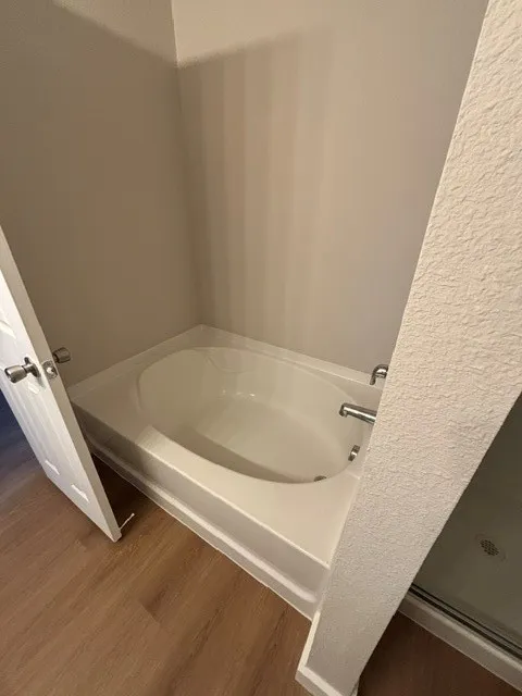 White drop-in bathtub with polished chrome fixtures, surrounded by light gray walls and wood-finish flooring