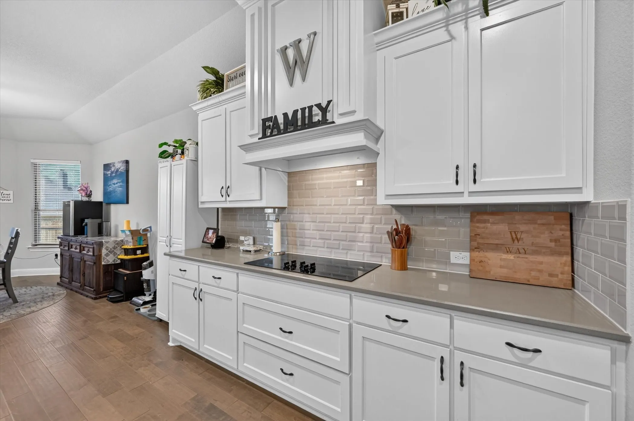 White cabinetry with dark hardware, a built-in cooktop, light-toned backsplash tile, and wood-finish flooring