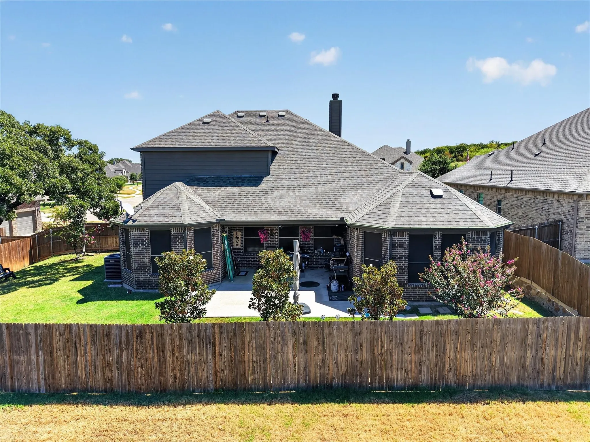 Exterior featuring a brick facade, shingle roof, privacy fence, green lawn, and a concrete patio