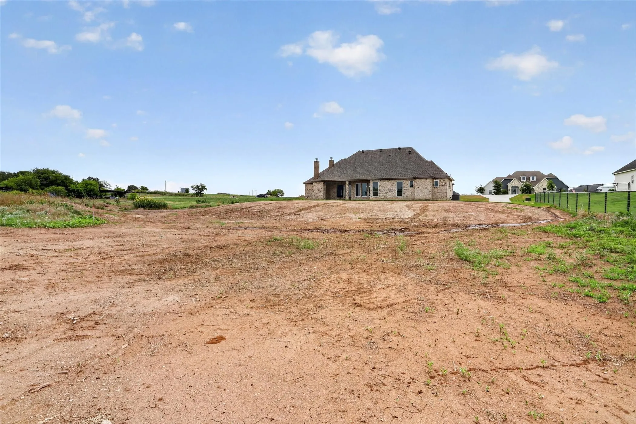 Large undeveloped lot featuring exposed earth, distant residential structures, and a partial perimeter fence