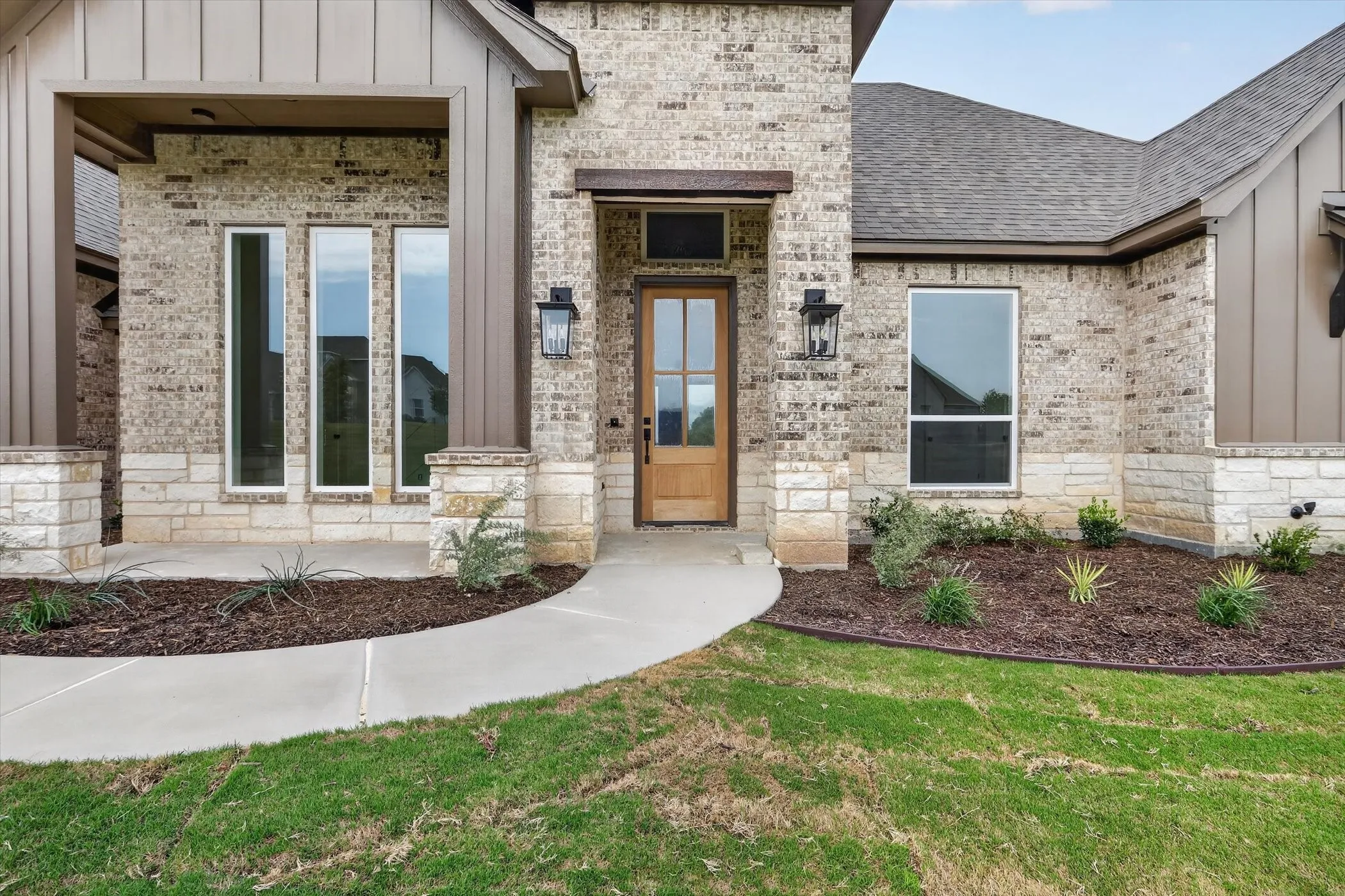 Brick and stone exterior featuring vertical panel siding accents, an inviting entryway with a wood-finish door, exterior wall lanterns, and a concrete pathway