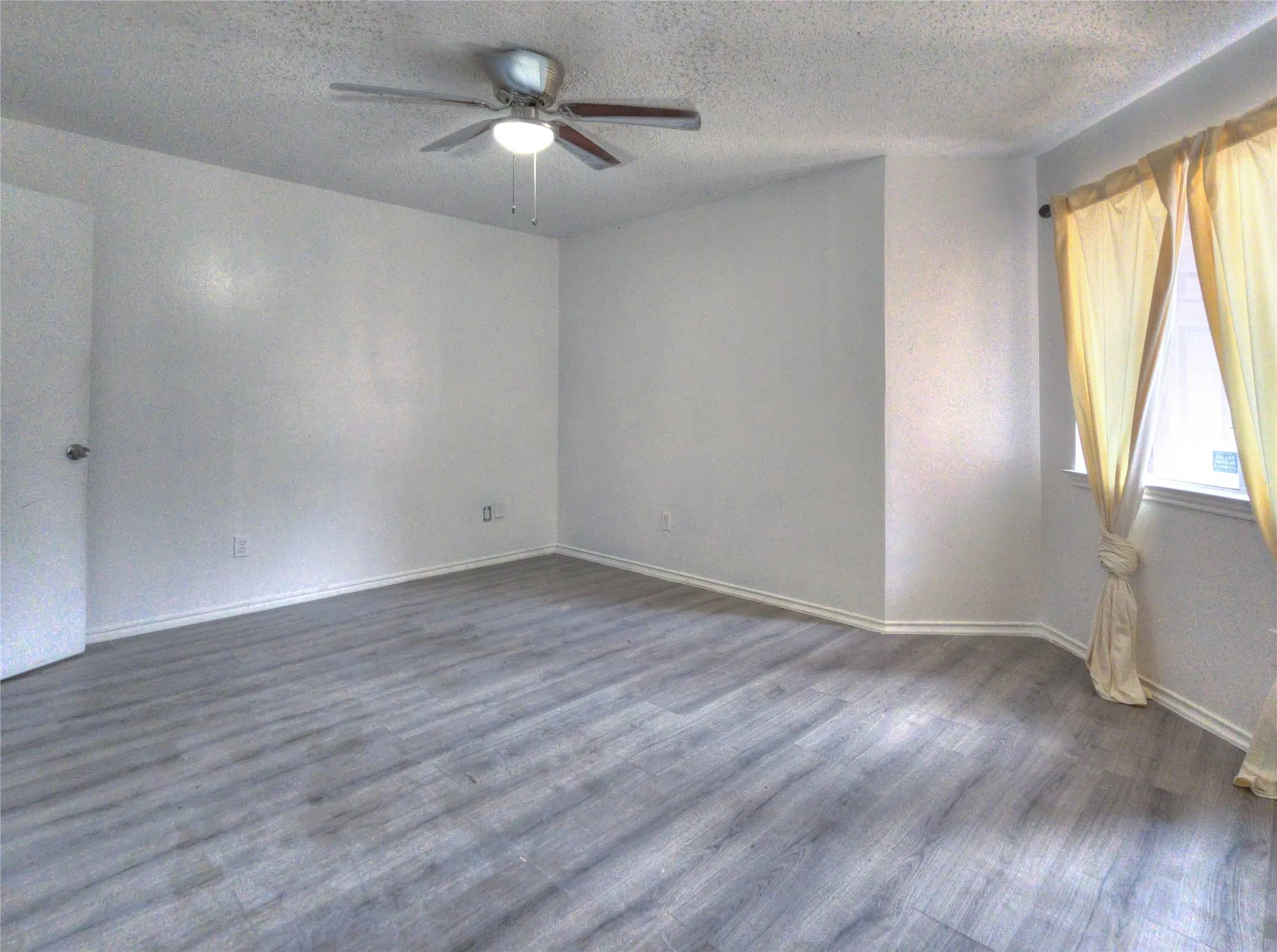 Spacious room featuring light gray wood-finish flooring, white walls, a ceiling fan with integrated lighting, and a prominent window