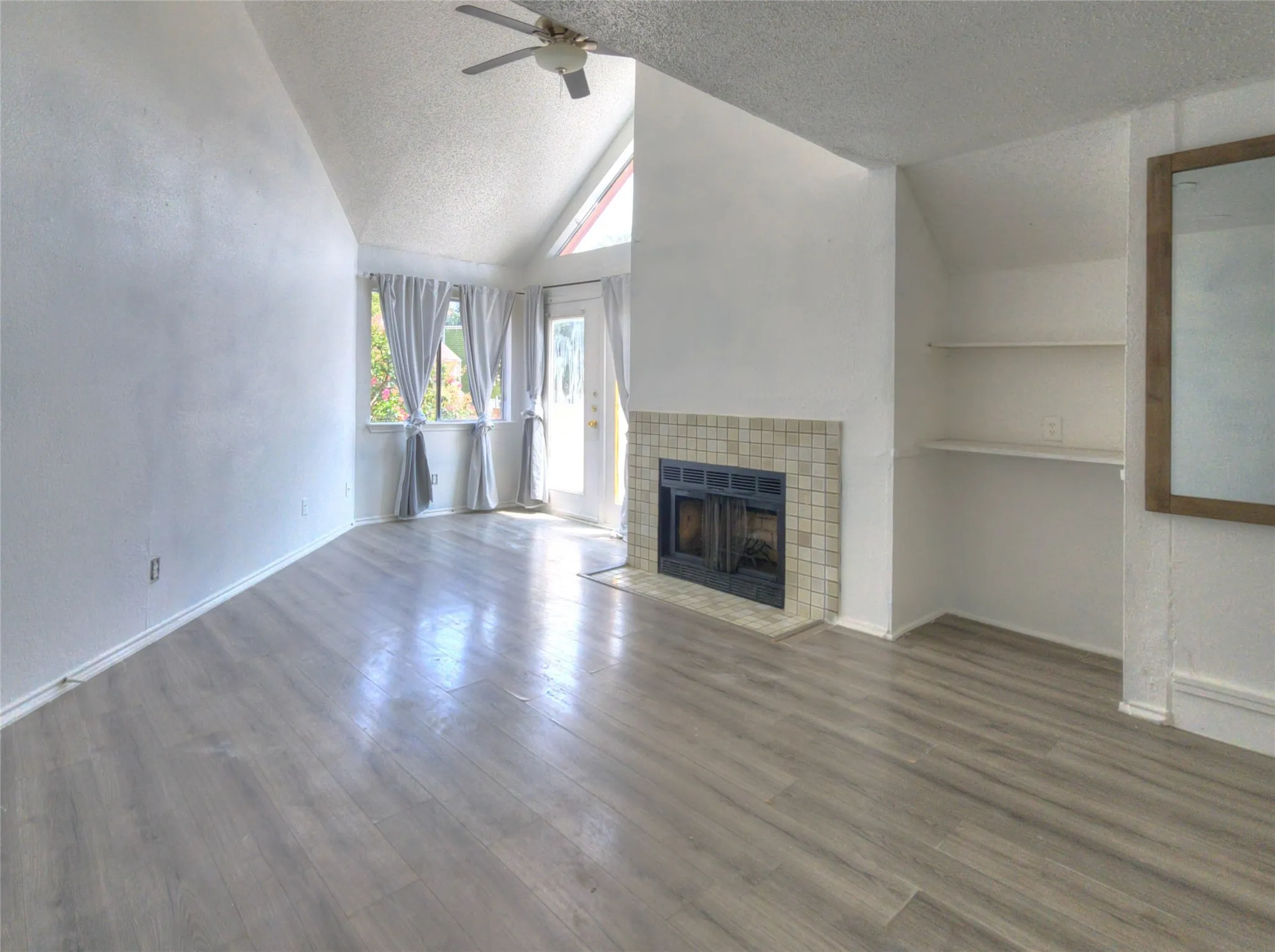 Vaulted ceiling with a ceiling fan, wood-finish flooring, fireplace with tile surround, built-in shelving, and exterior access via a glass-paneled door