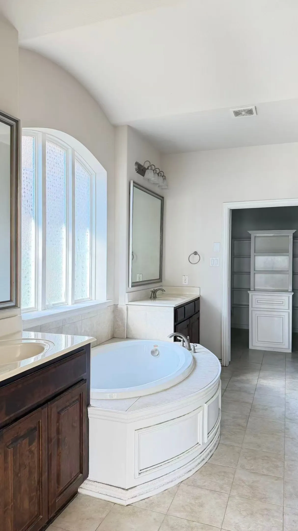 Built-in soaking tub with decorative paneling, arched window, dual vanity sinks, and tile flooring
