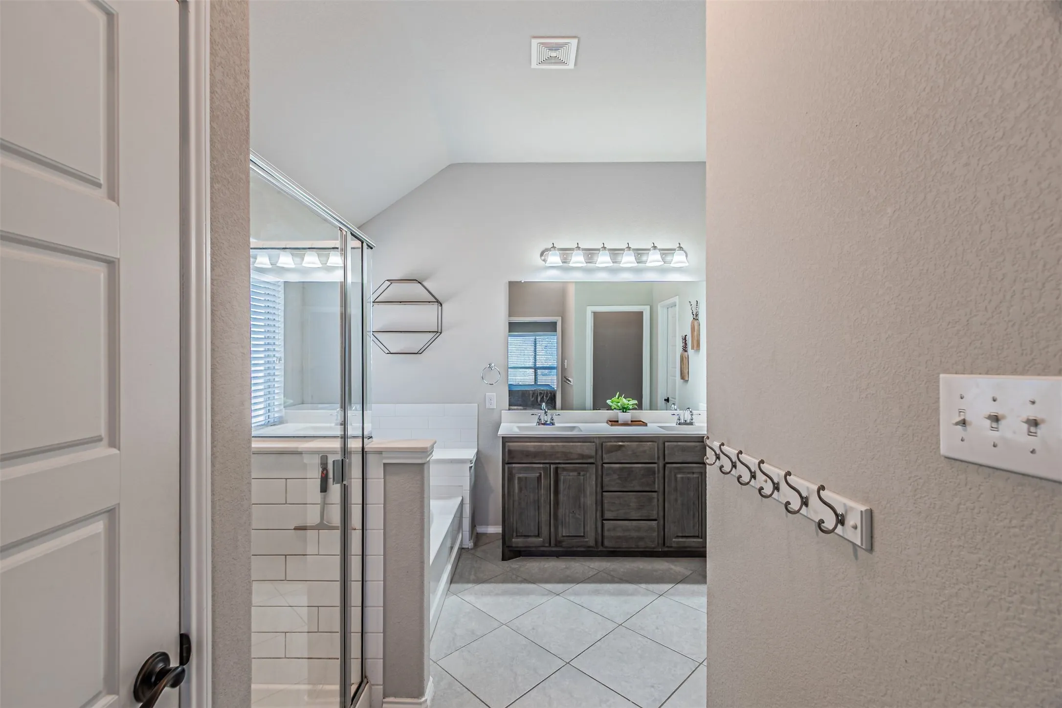 Full bath featuring double vanity, a textured wall, a shower stall, light tile patterned floors, and lofted ceiling