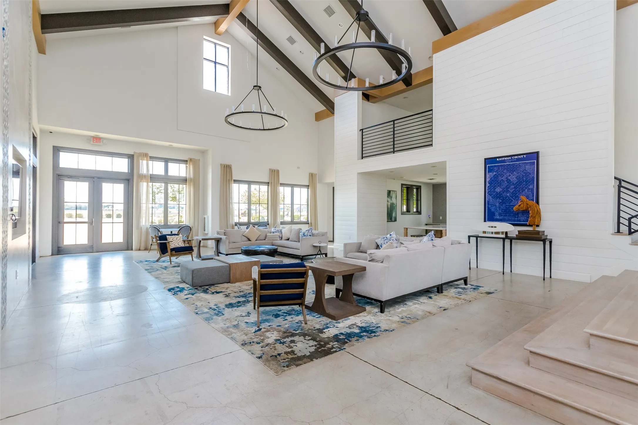 Soaring vaulted ceilings with exposed wood beams, dual circular chandeliers, extensive window banks, and a second-story balcony with horizontal bar railing