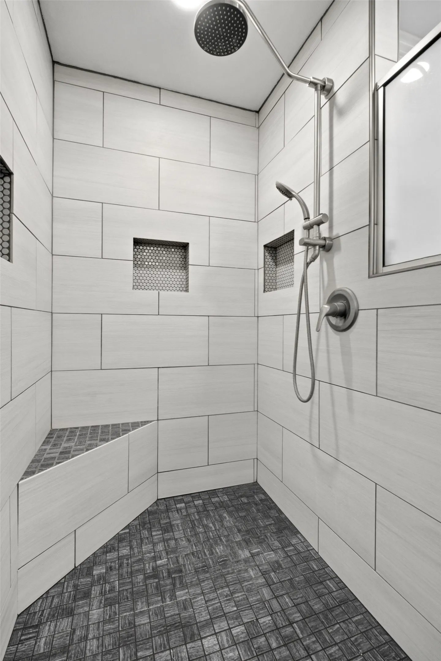 Walk-in shower with large format wall tiling, a built-in bench, two recessed niches, a rain shower head, and a handheld shower​​‌​​​​‌​​‌‌​‌‌​​​‌‌​‌​‌​‌​​​‌​​ fixture