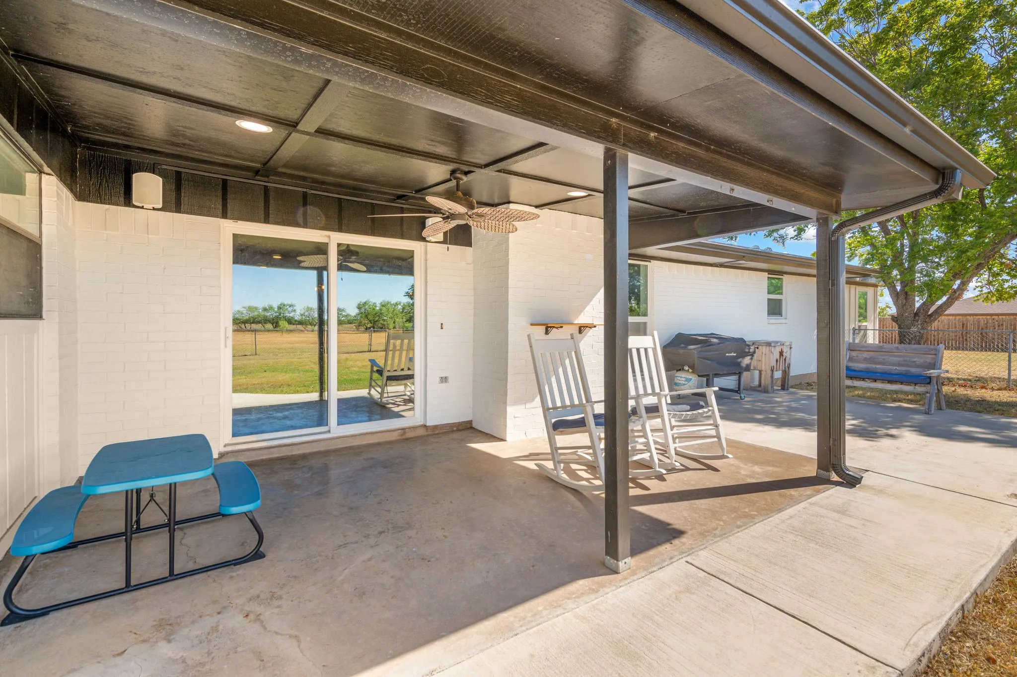 Covered patio with concrete flooring, supported by dark metal posts, featuring a ceiling fan and recessed lighting