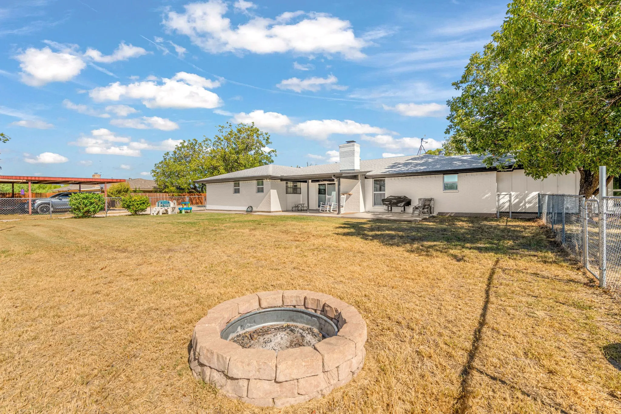 Expansive backyard featuring a stone-clad fire pit, mature trees, and a covered patio area