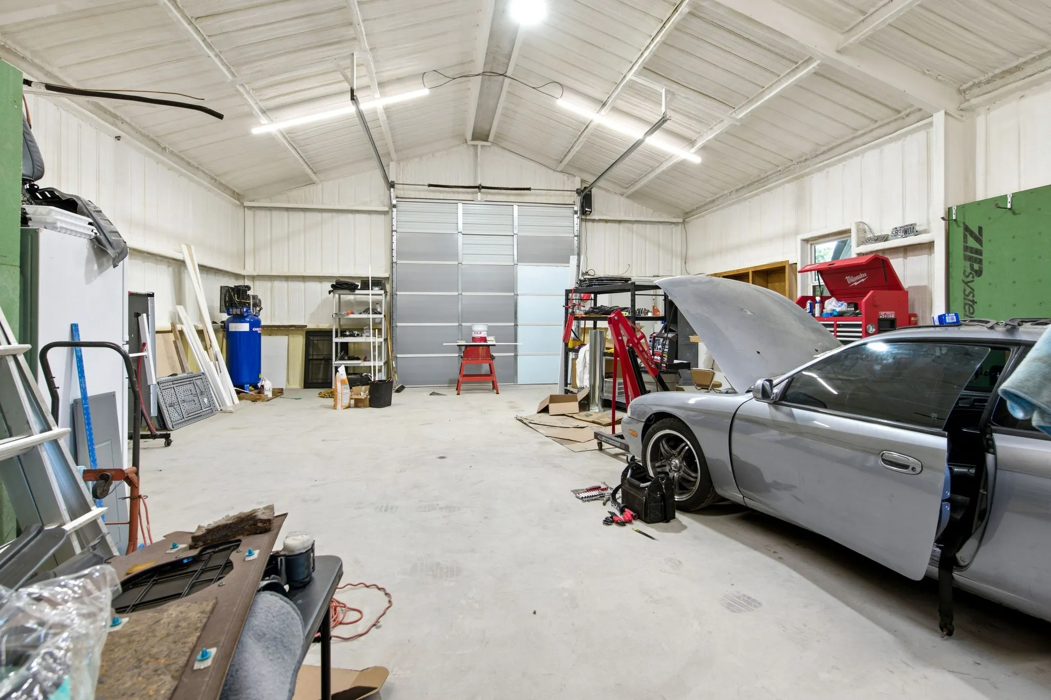 Spacious Workshop with a large roll-up door, exposed metal beam ceiling, concrete flooring, and ample overhead lighting