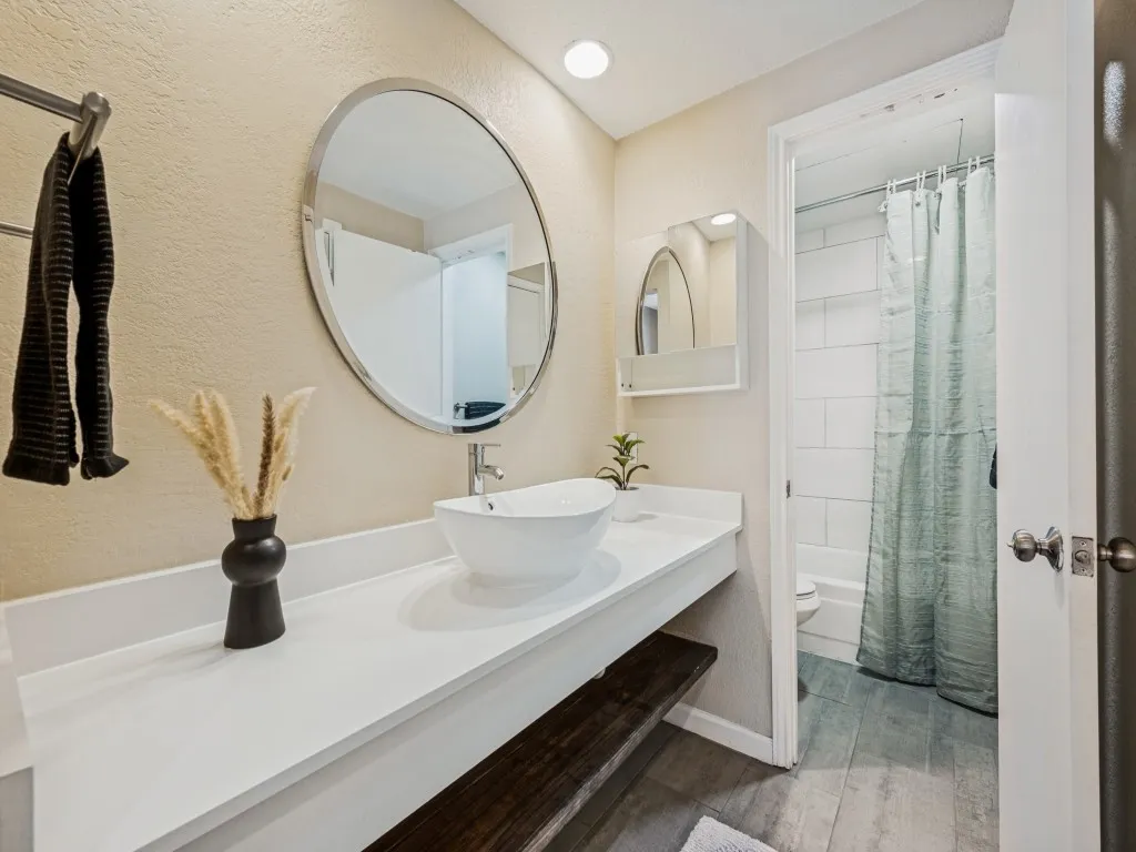 Floating vanity with a vessel sink, a large circular mirror, wood-finish flooring, and a white tiled shower with a bathtub