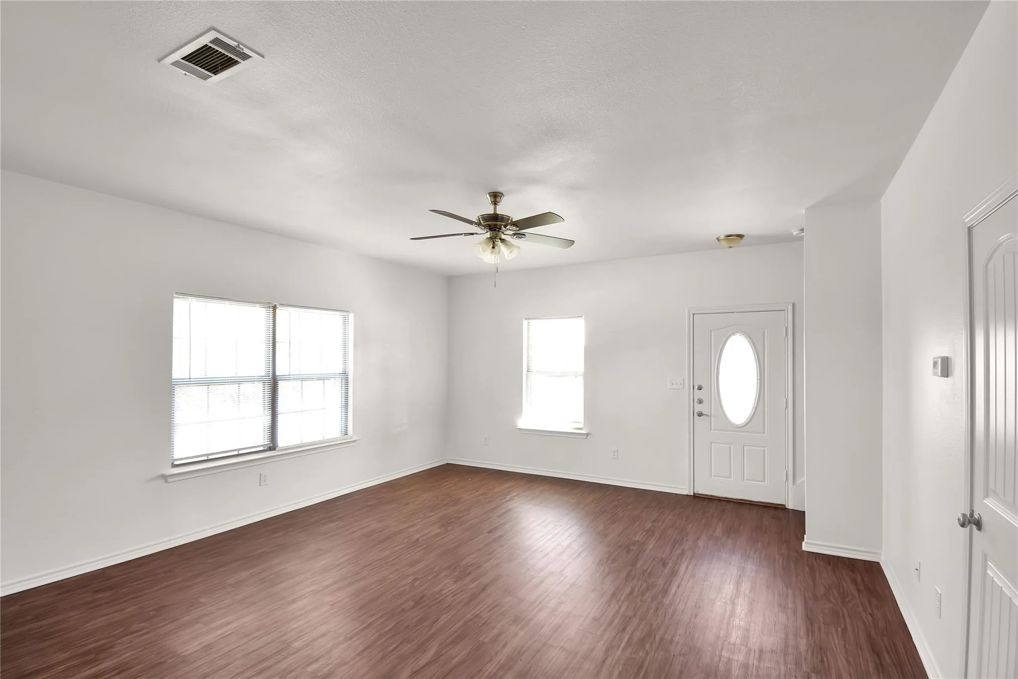 Wood-finish flooring, white walls, multiple windows, ceiling fan with integrated lighting, and a white front door with an oval glass insert
