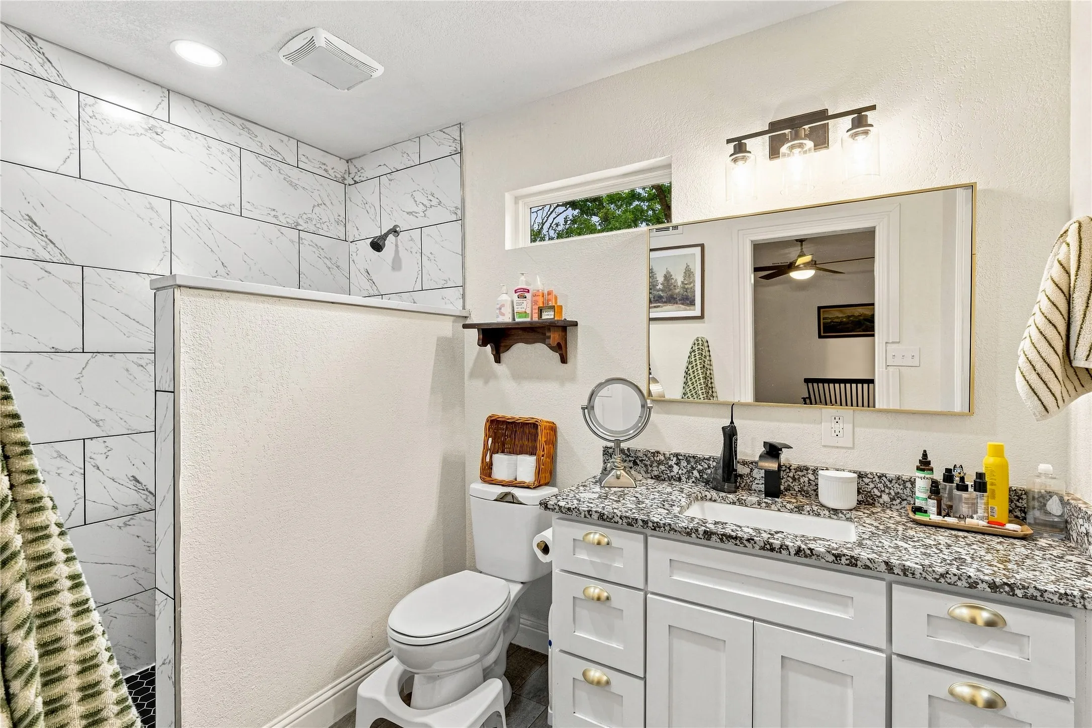 Walk-in shower with large format tile, a single vanity with granite countertop, white cabinetry with gold-tone hardware, and an attached rectangular mirror
