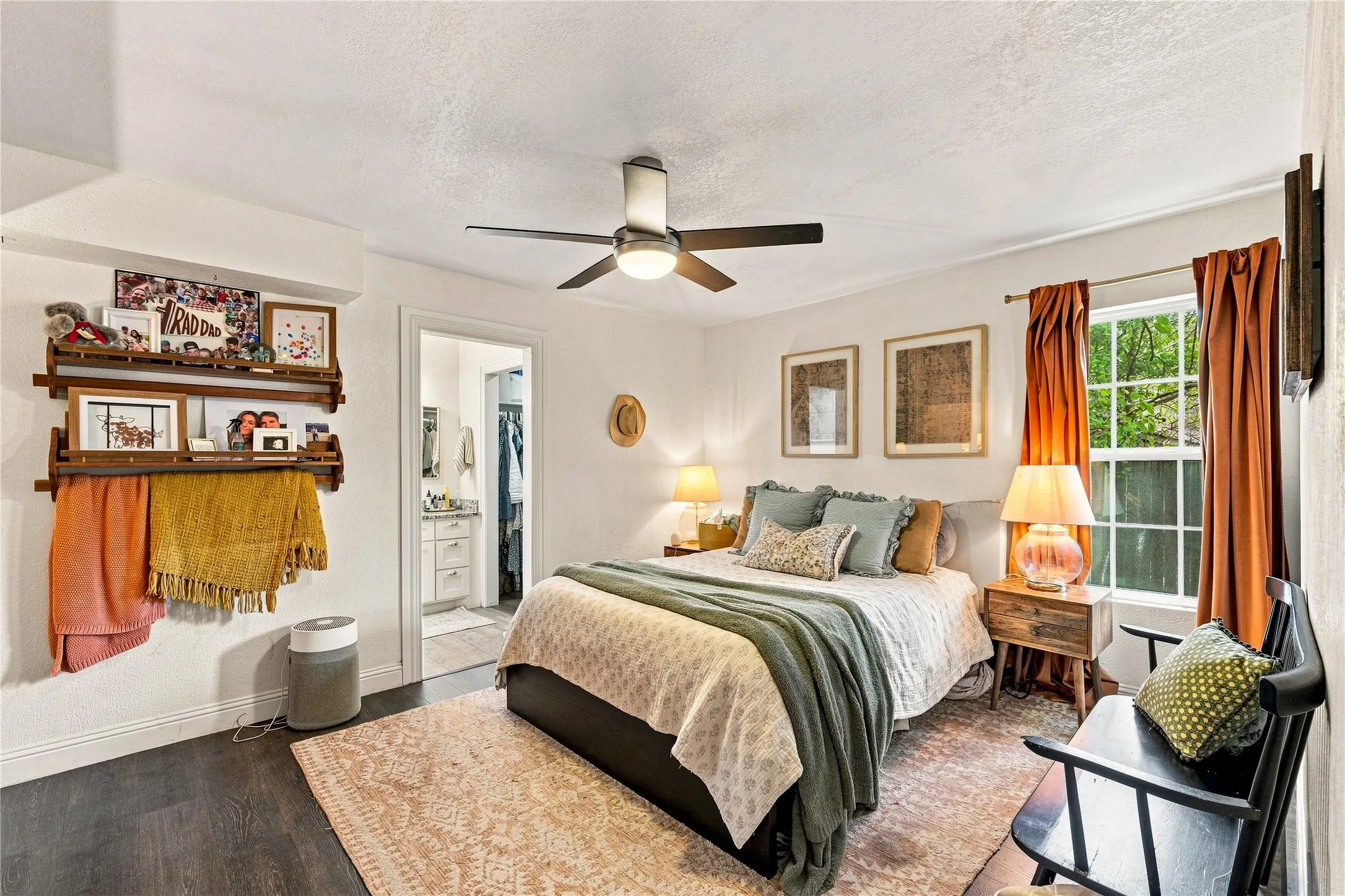 Dark wood-finish flooring, ceiling fan with integrated lighting, wood wall shelving, and an en-suite bathroom with a vanity