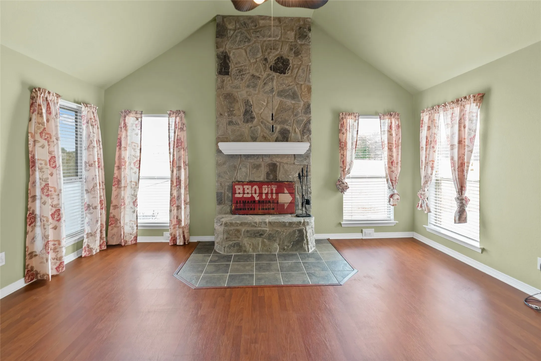 Vaulted ceiling with a ceiling fan, stone-clad fireplace with a mantel, tile hearth, and wood-finish flooring