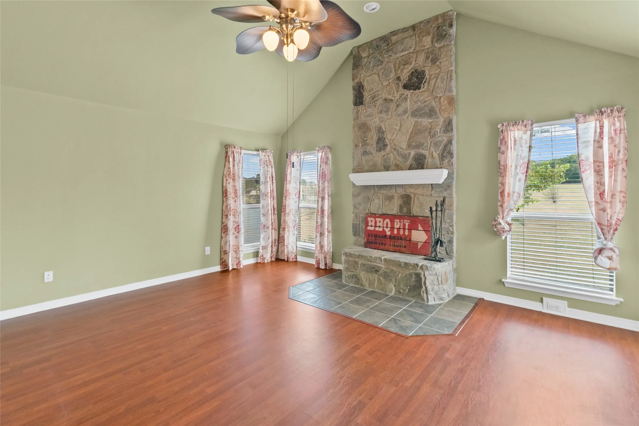 Vaulted ceiling, wood-finish flooring, stone-clad fireplace with mantel, tiled hearth, and multiple windows