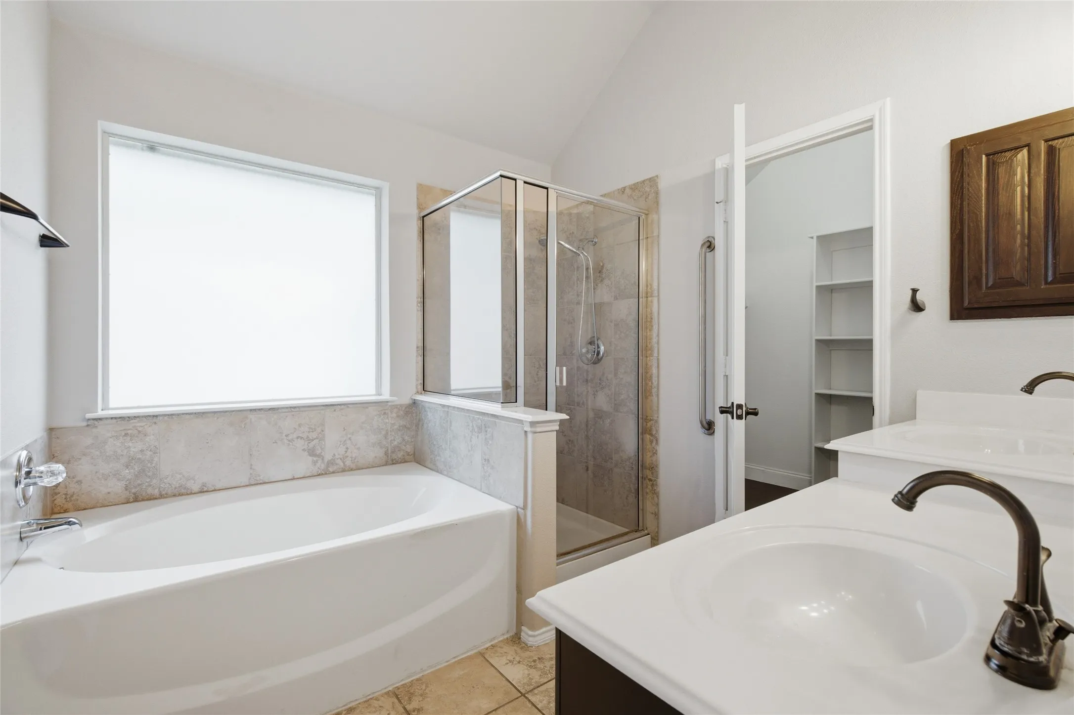 Dual vanity with undermount sinks and bronze-finish faucets, built-in soaking tub with tile surround, glass-enclosed shower with tile, and a vaulted ceiling
