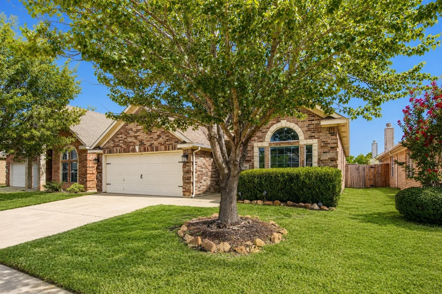 Brick exterior with an arched window, two-car garage, concrete driveway, and mature​​‌​​​​‌​​‌‌​‌‌​​​‌‌​‌​‌​‌​​​‌​​ landscaping
