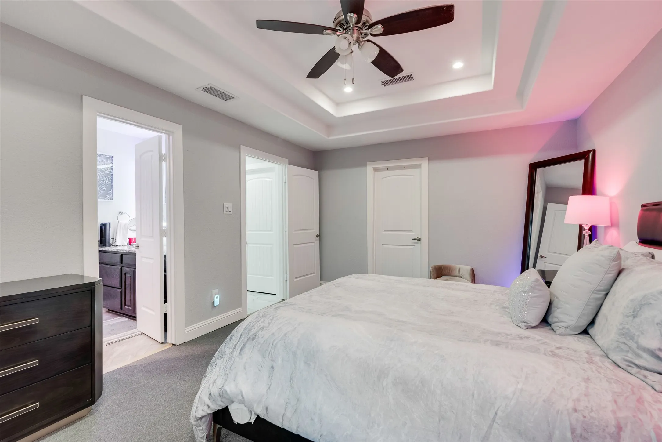 Carpeted bedroom with a tray ceiling, baseboards, ensuite bathroom, recessed lighting, and a ceiling fan