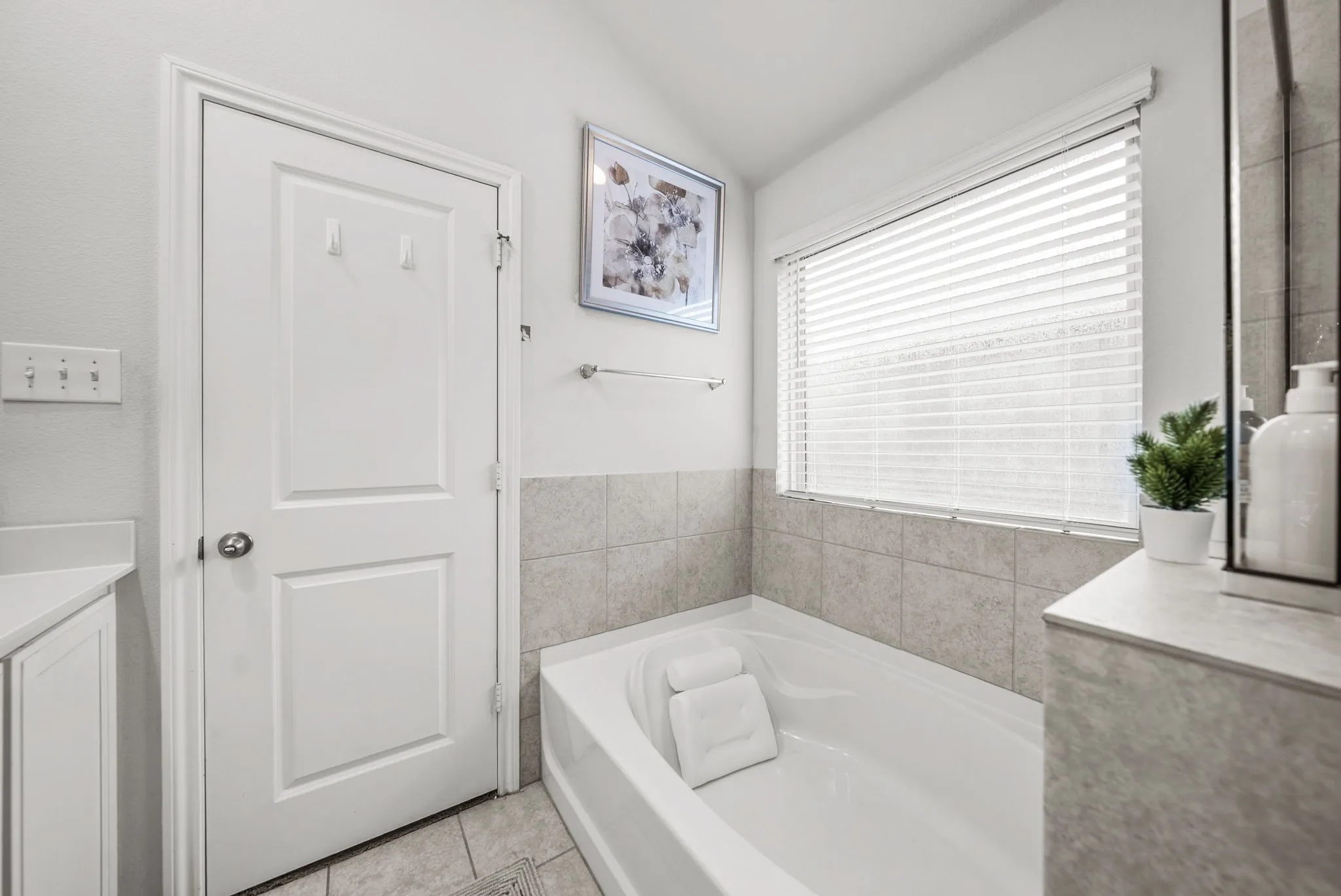 Bathtub with tiled surround, light-colored wall paint, and a window with blinds