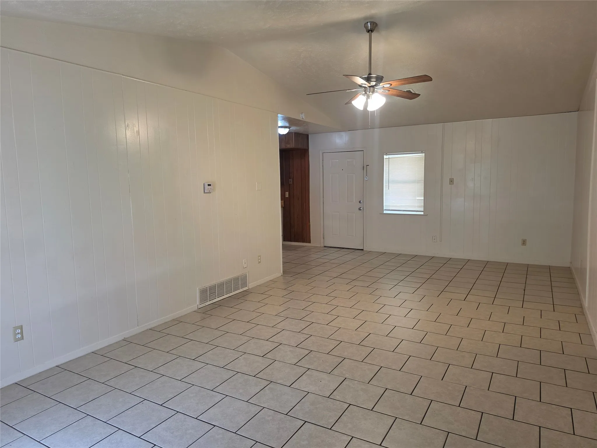 Spacious living area featuring light-colored tile flooring, white paneling, a ceiling fan with integrated lighting, and a white entry door