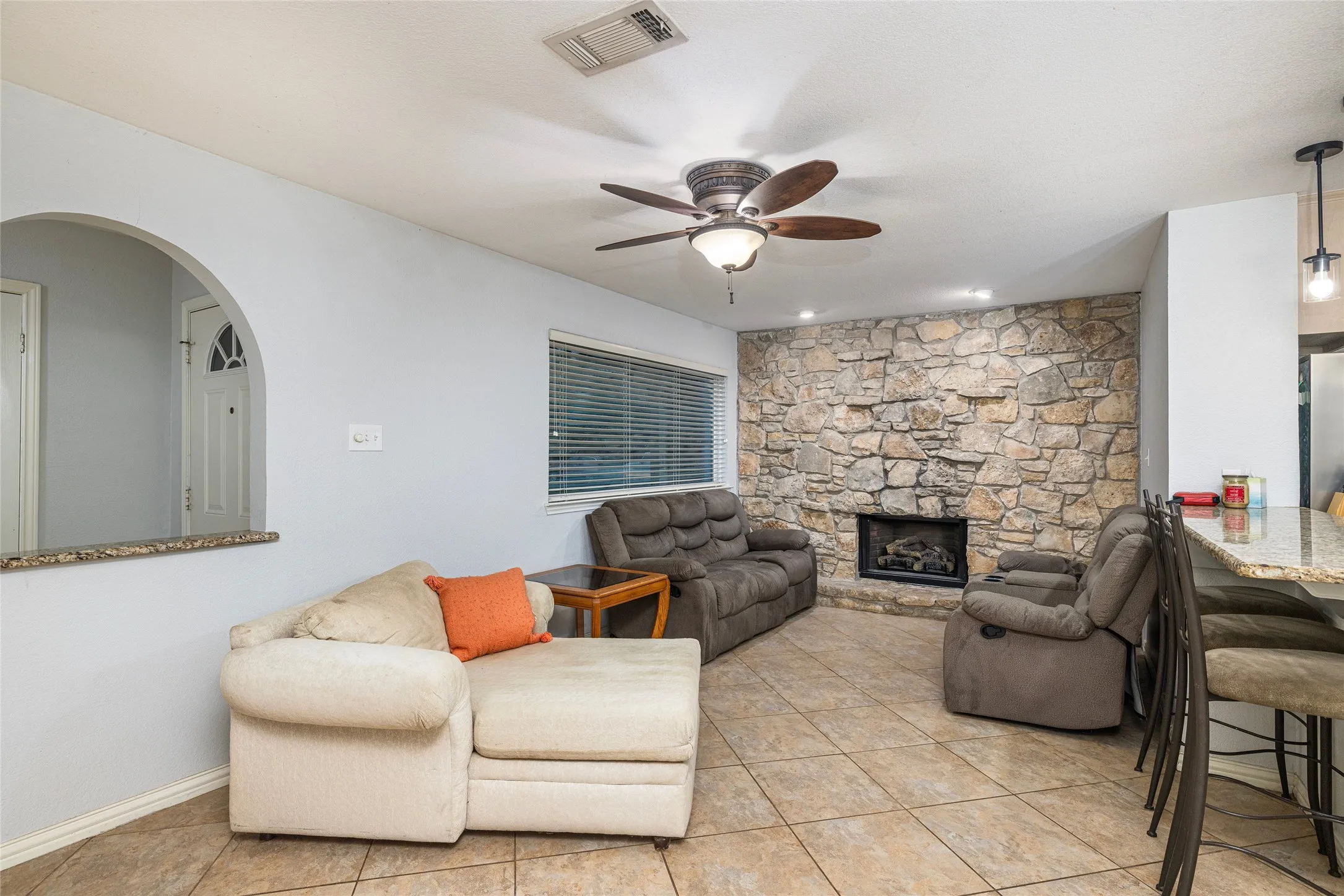 Living area featuring a stone accent wall with a fireplace, a ceiling fan, tiled flooring, and a breakfast bar with a granite-style countertop